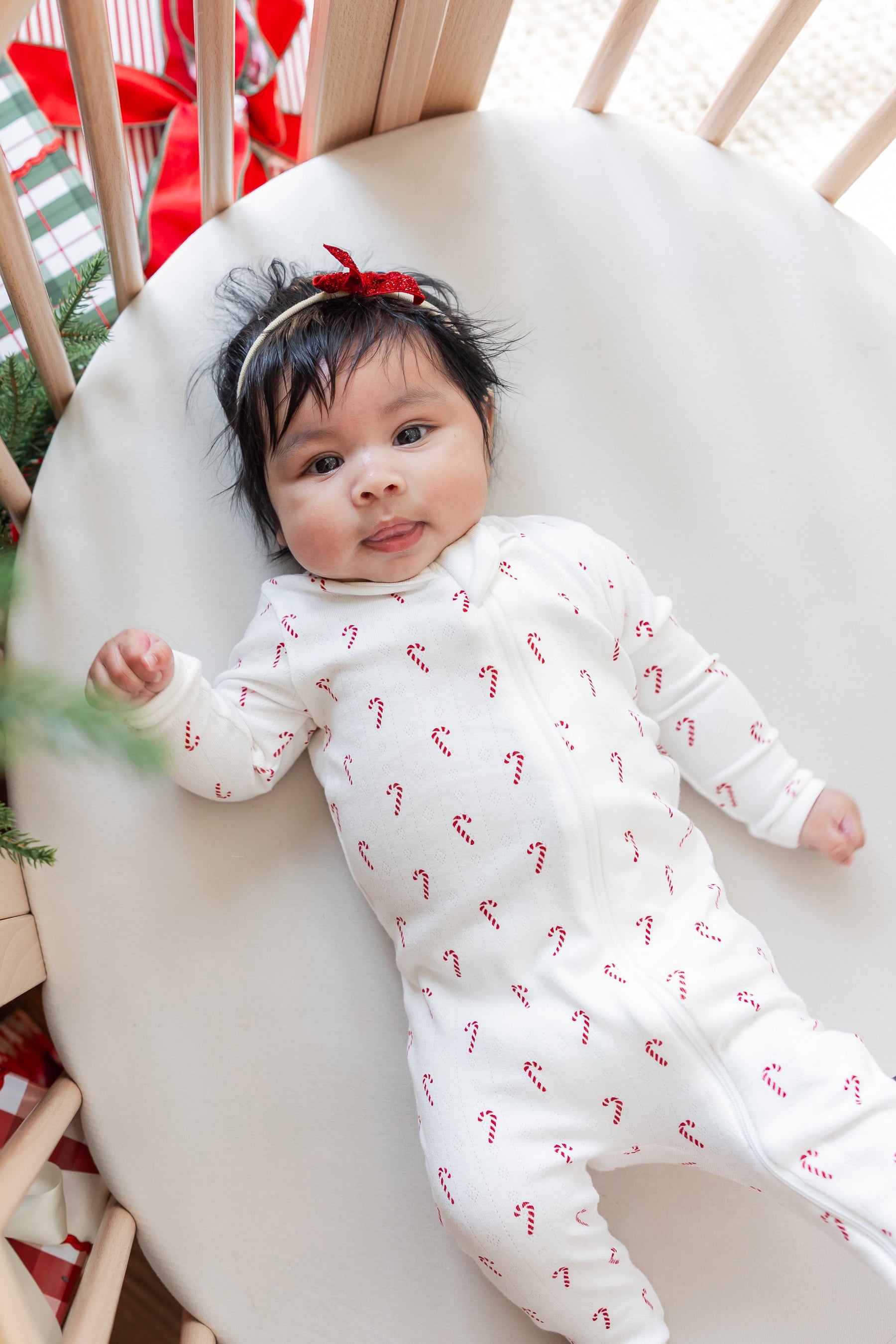 Baby wearing Pointelle Zippered Footie in Candy Cane and bow headband lying in a crib near a Christmas tree