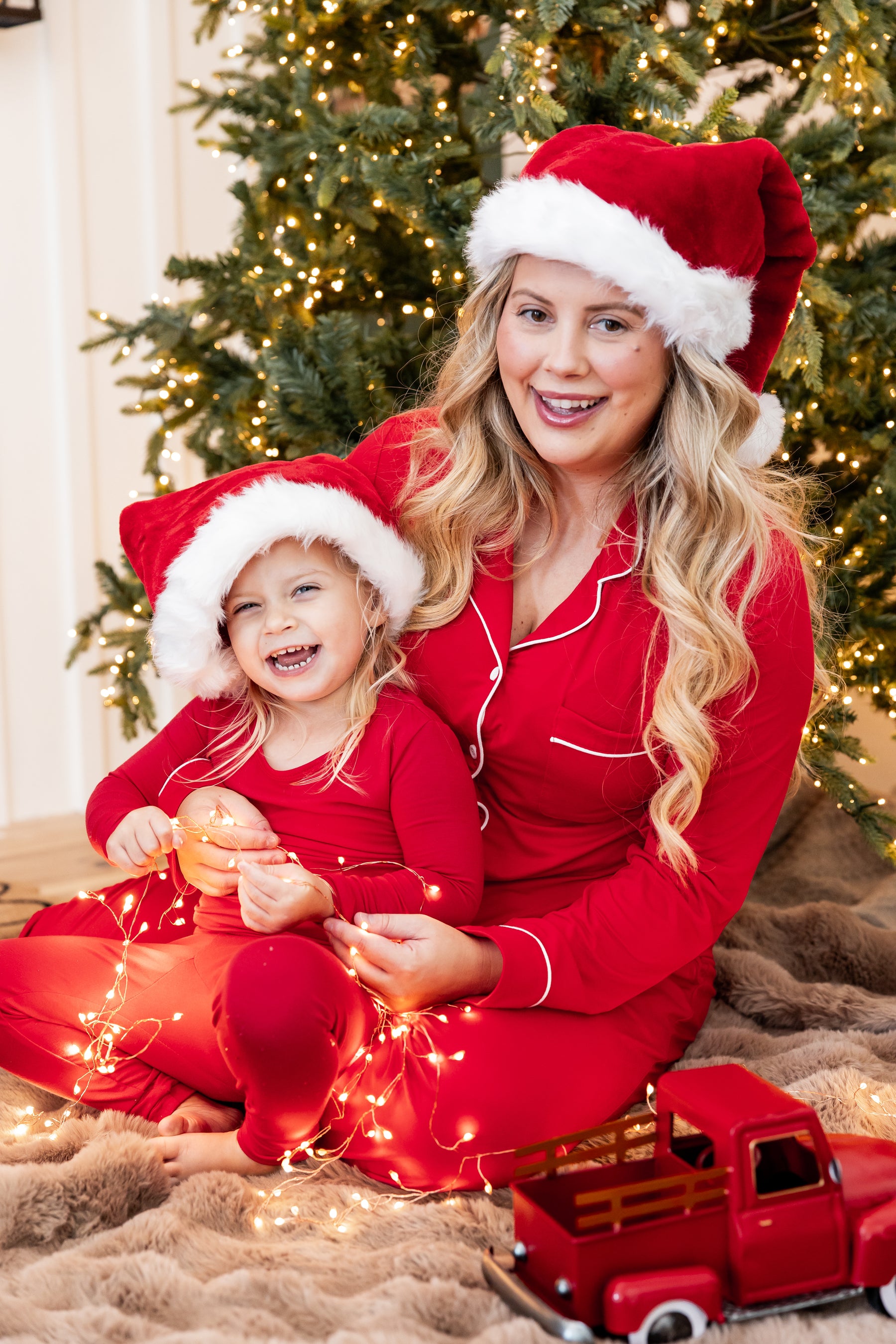 A woman and a young girl wearing matching red pajamas and Santa hats sit together on a brown faux-fur rug in front of a decorated Christmas tree, with warm string lights and a small red toy truck around them.