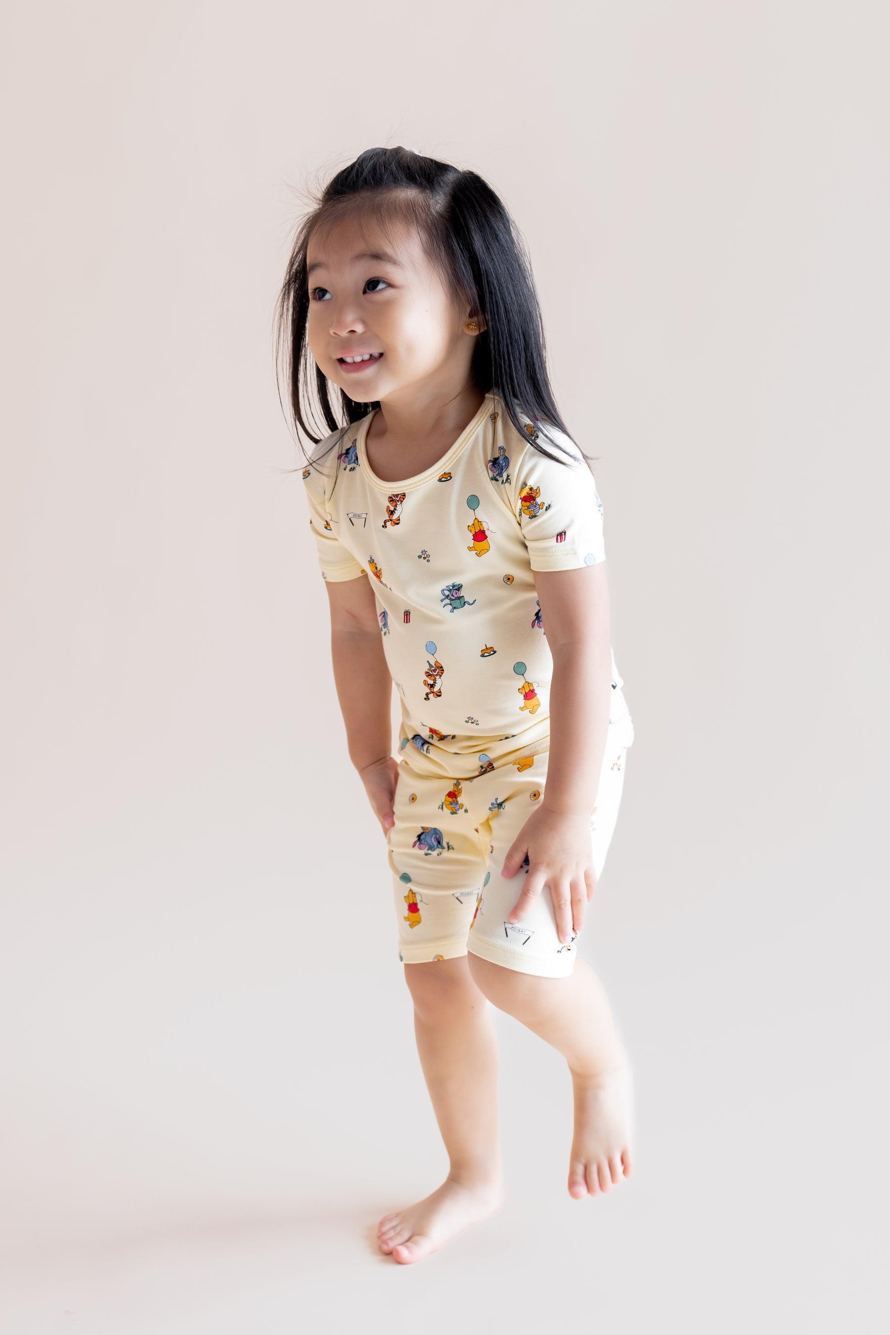Girl standing wearing Short Sleeve Pajamas in Winnie the Pooh Celebration print on a cream background