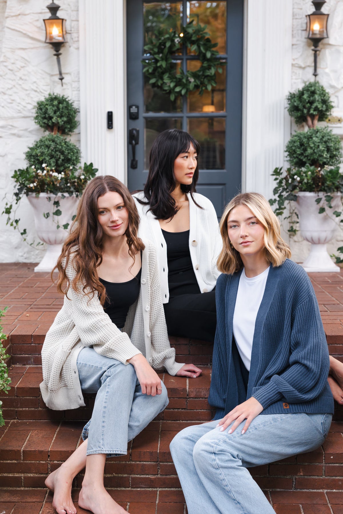 Three modal sitting outdoors wearing Chunky Knit Women's Cropped Cardigan in Cloud, Chunky knit women's waffle cardigan in almond and chunky knit oversized cardigan in oxford