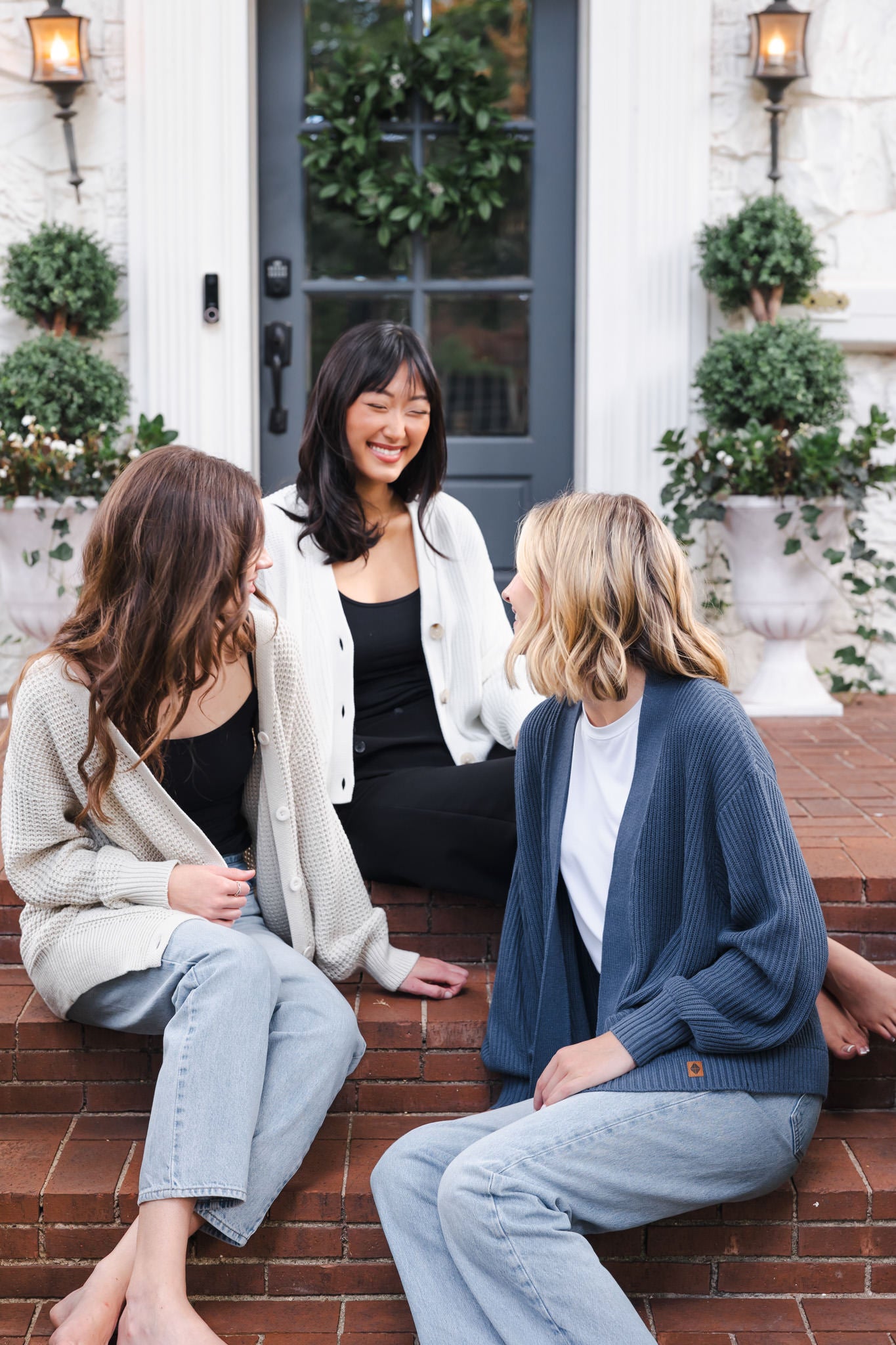 Three modal sitting outdoors wearing Chunky Knit Women's Cropped Cardigan in Cloud, Chunky knit women's waffle cardigan in almond and chunky knit oversized cardigan in oxford