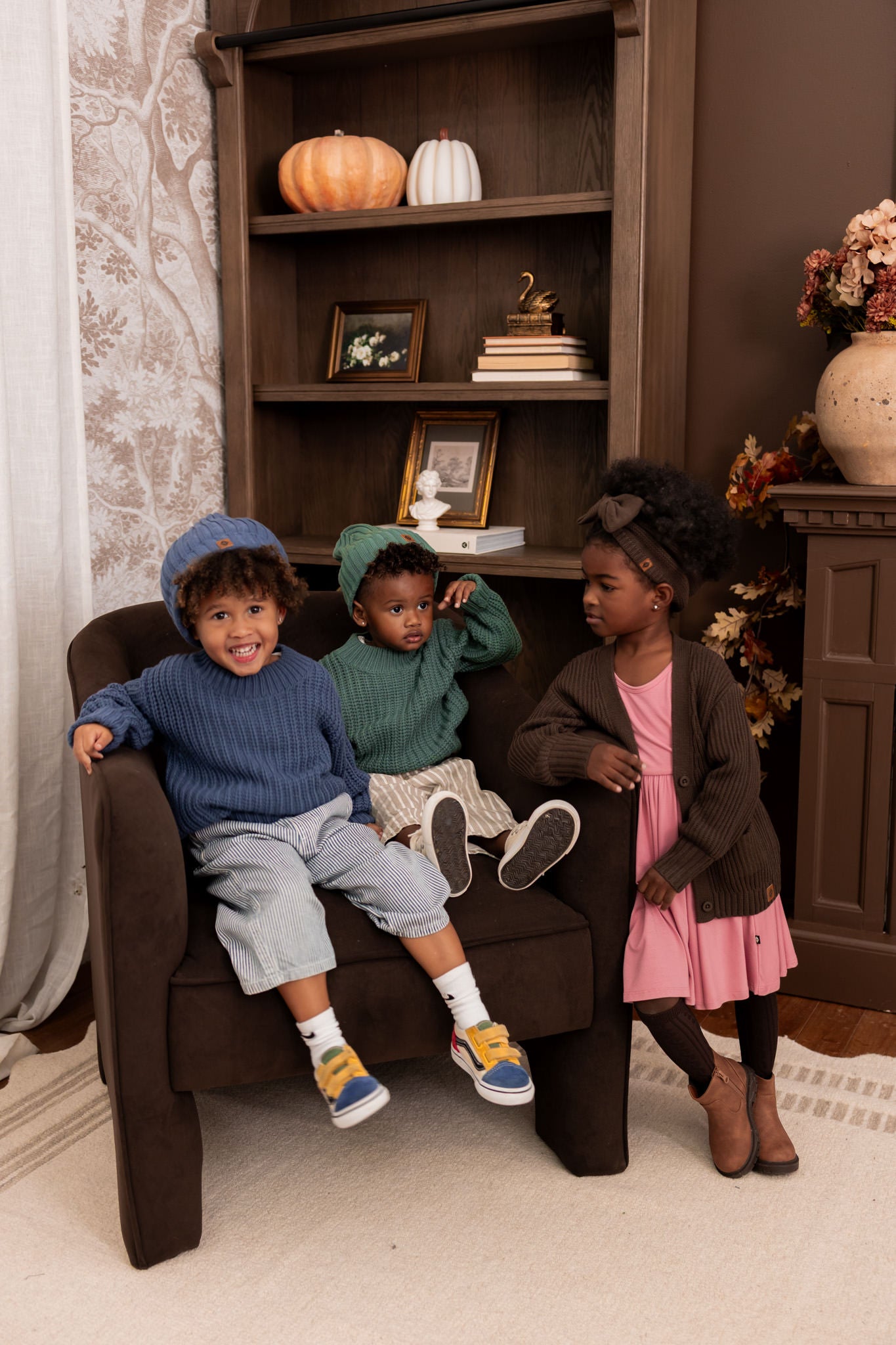 Two boys sitting on sofa wearing Chunky Knit Sweater in Pine and oxford with chunky knit ribbed beanies in pine and oxford with a girl standing beside the sofa wearing chunky knit oversized cardigan in espresso