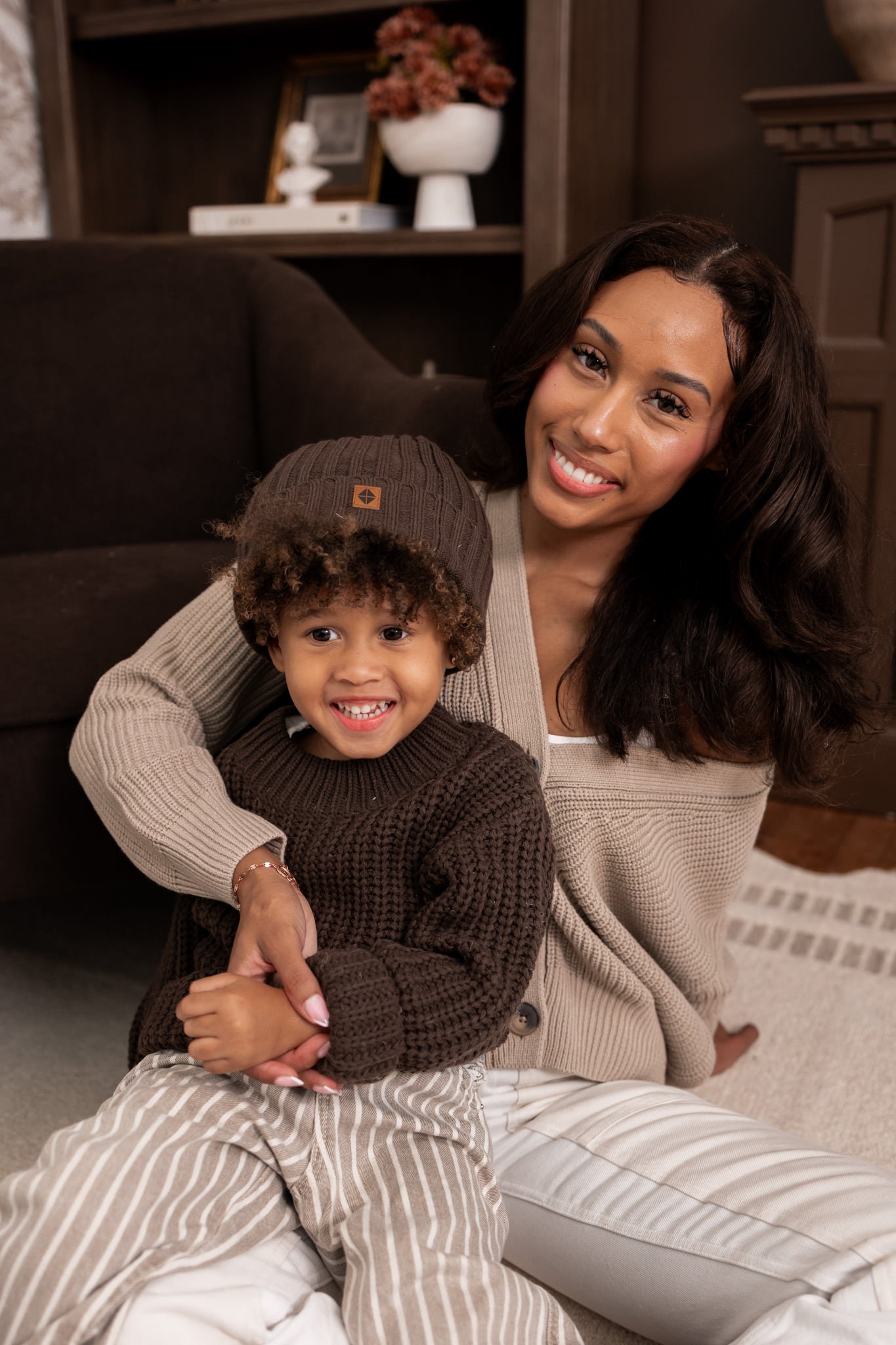 Modal smiling in Chunky Knit Women's Cropped Cardigan in Almond with child wearing chunky knit ribbed beanie and Chunky knit sweater in espresso sitting in al living room