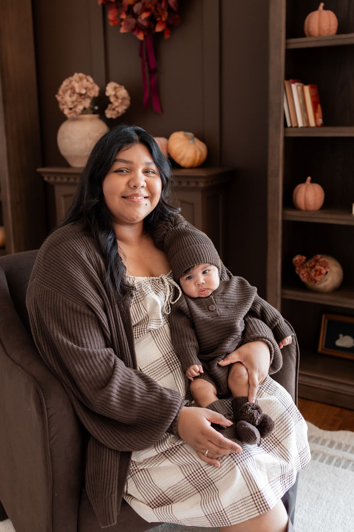 Mommy posing on brown sofa wearing Chunky Knit Women's Oversized Cardigan in Espresso holding a baby wearing Chunky knit romper, chunky knit pom beanie and knit booties in espresso