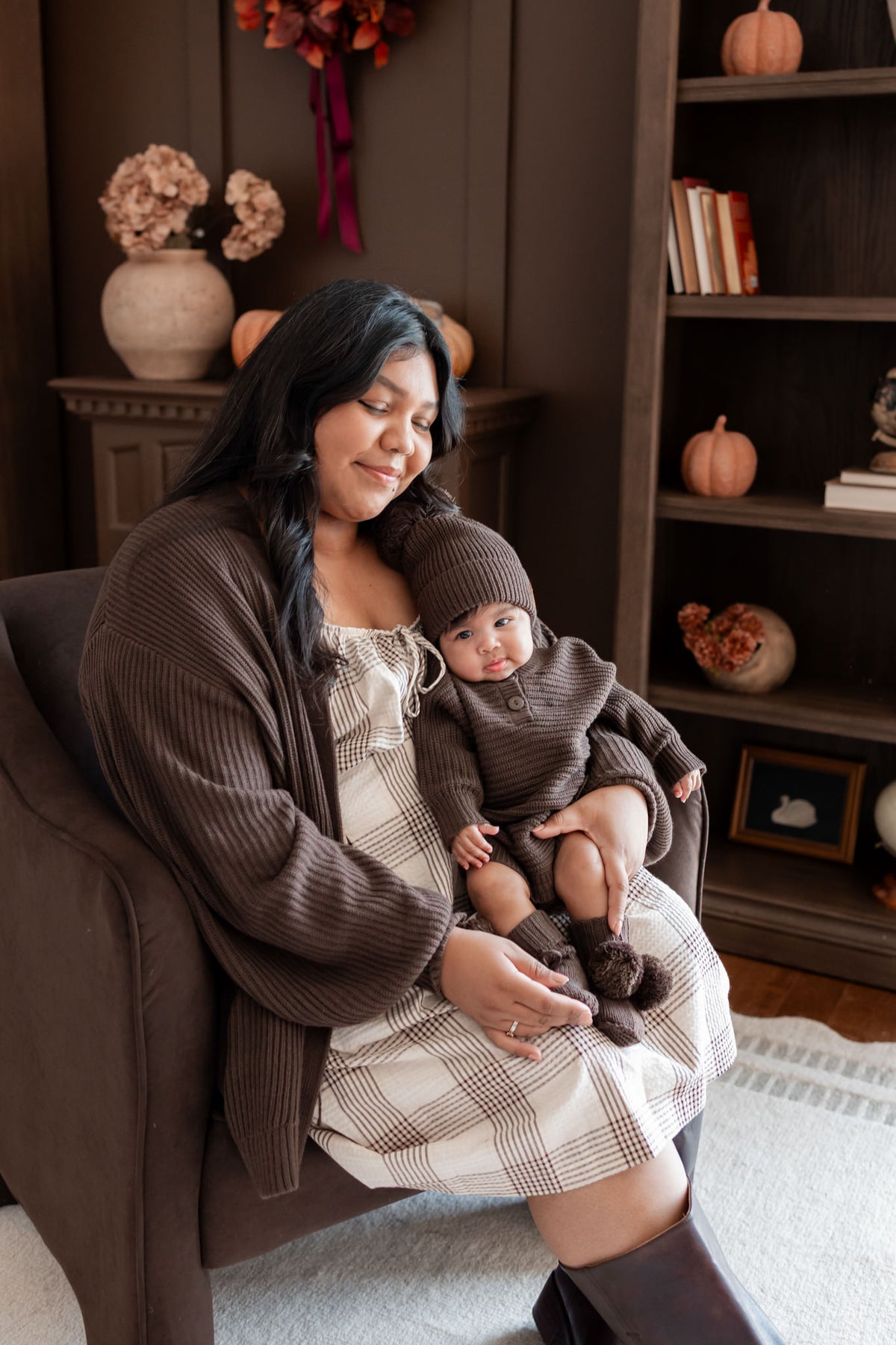 Mommy sitting on brown sofa wearing Chunky Knit Women's Oversized Cardigan in Espresso holding a baby wearing Chunky knit romper, chunky knit pom beanie and knit booties in espresso