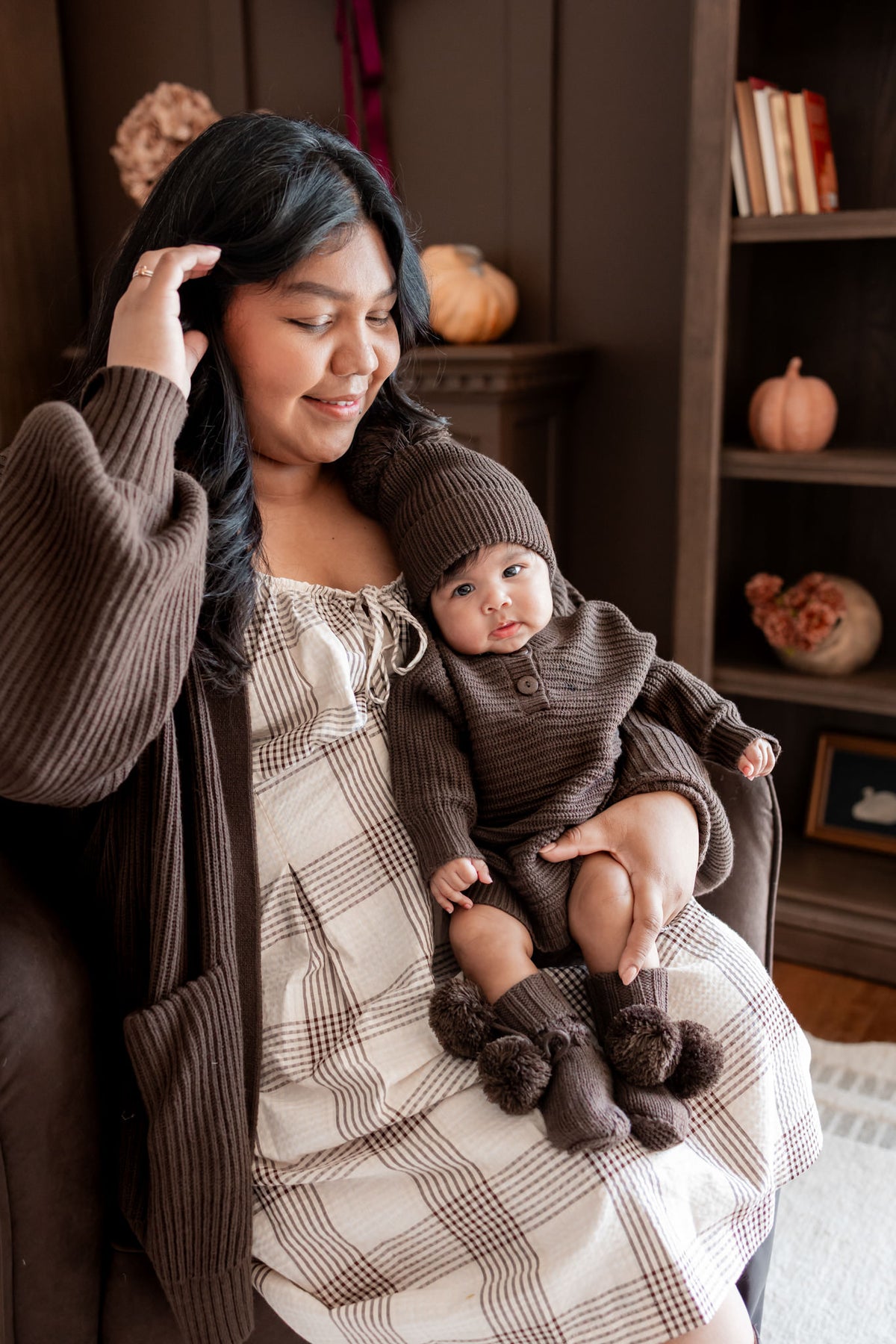 Mommy sitting on brown sofa wearing Chunky Knit Women's Oversized Cardigan in Espresso holding a baby wearing Chunky knit romper, chunky knit pom beanie and knit booties in espresso