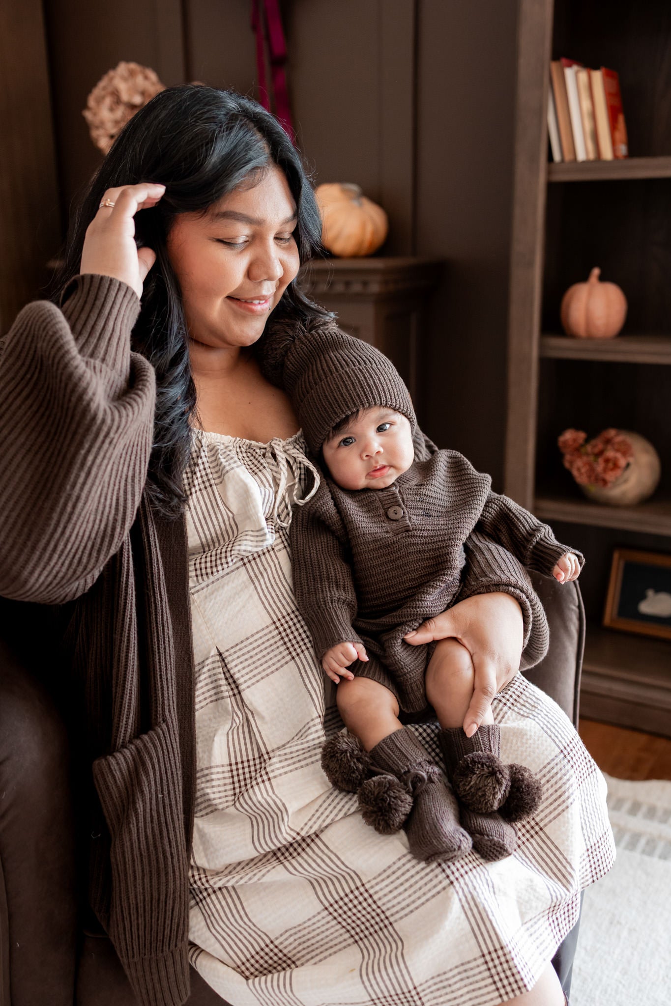 Mommy sitting on brown sofa wearing Chunky Knit Women's Oversized Cardigan in Espresso holding a baby wearing Chunky knit romper, chunky knit pom beanie and knit booties in espresso