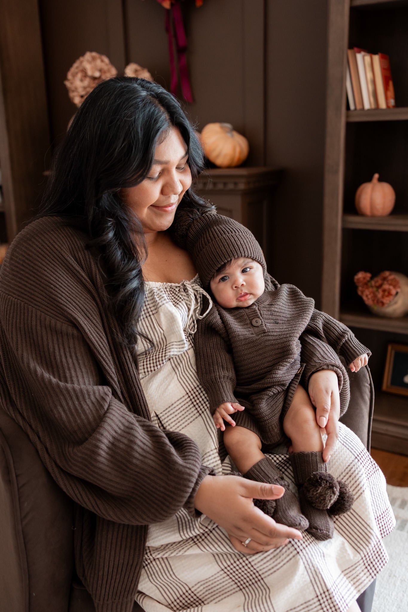 Mommy sitting on brown sofa wearing Chunky Knit Women's Oversized Cardigan in Espresso holding a baby wearing Chunky knit romper, chunky knit pom beanie and knit booties in espresso