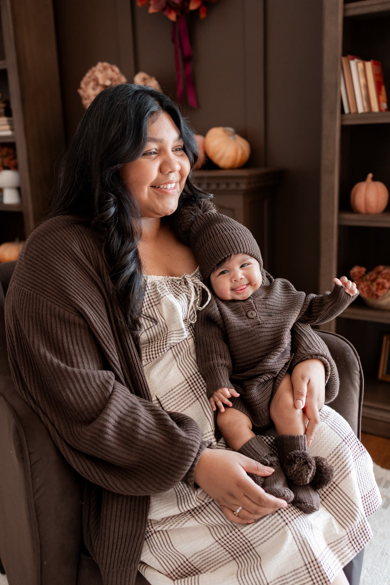 Mommy sitting on brown sofa wearing Chunky Knit Women's Oversized Cardigan in Espresso holding a baby wearing Chunky knit romper, chunky knit pom beanie and knit booties in espresso smiling together in a living room