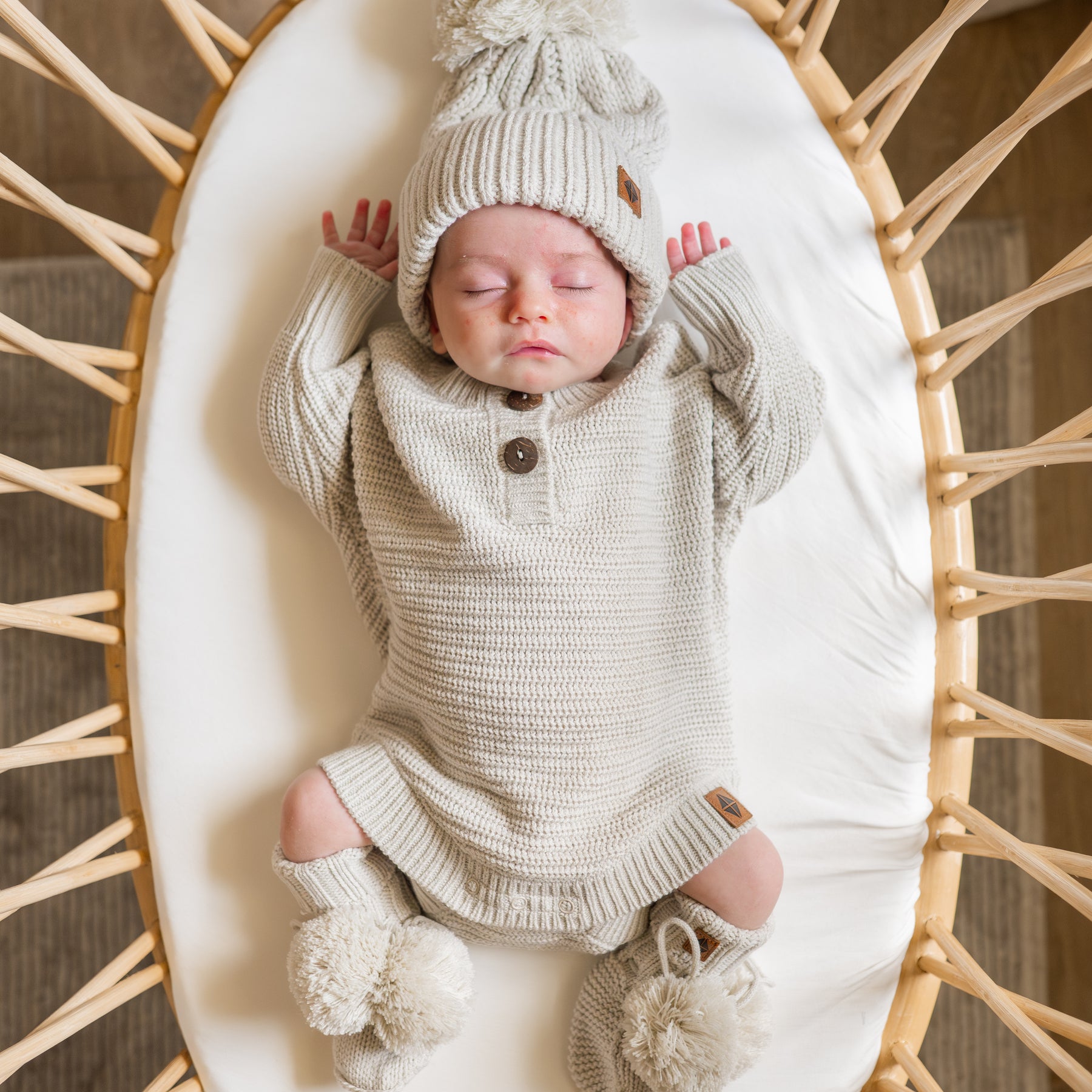 Sleeping newborn laying in a mini crib sheet wearing the Chunky Knit Romper in Oat and matching Chunky knit cable beanie and knit booties
