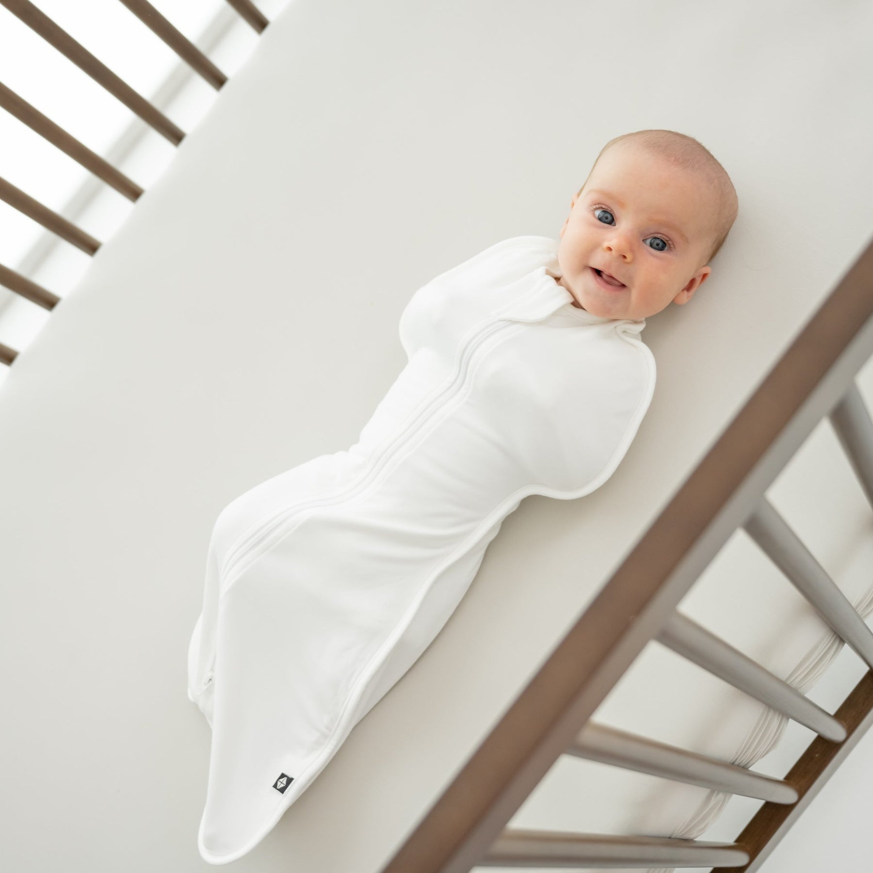 Baby in a wooden crib wearing a white swaddle bag 