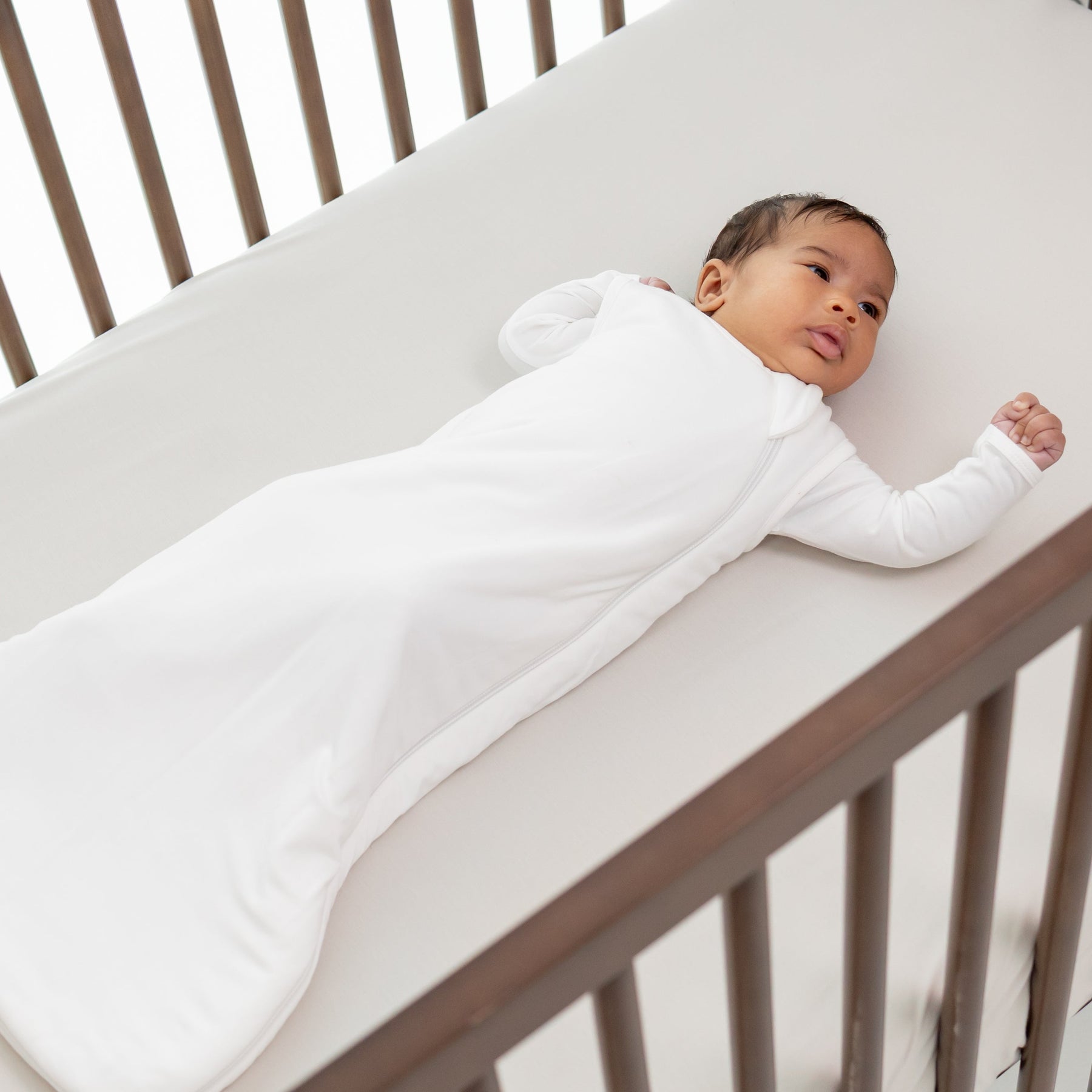 Baby in a small white sleep sack laying in a wooden crib