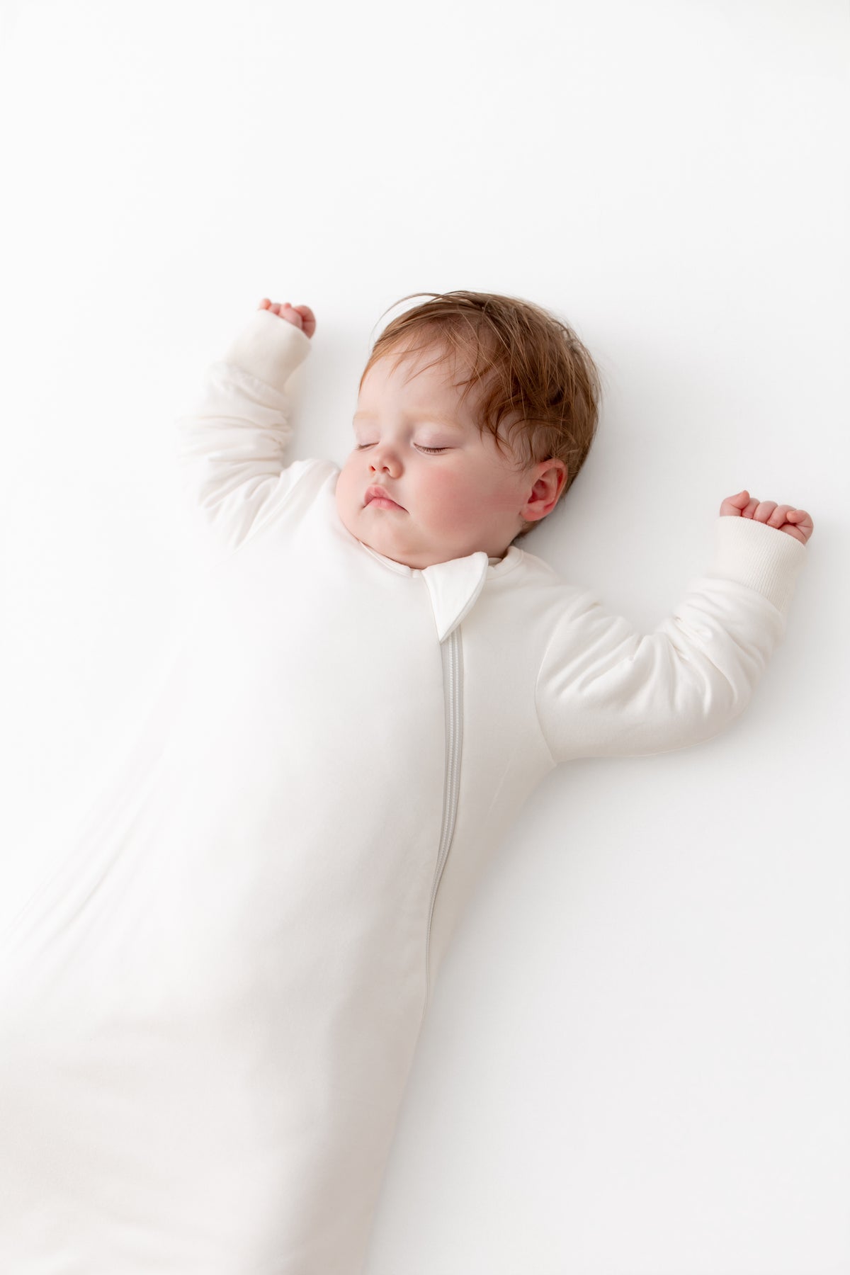 A sleeping baby lying on their back wearing a white long-sleeve zip-up sleeper against a clean white background.