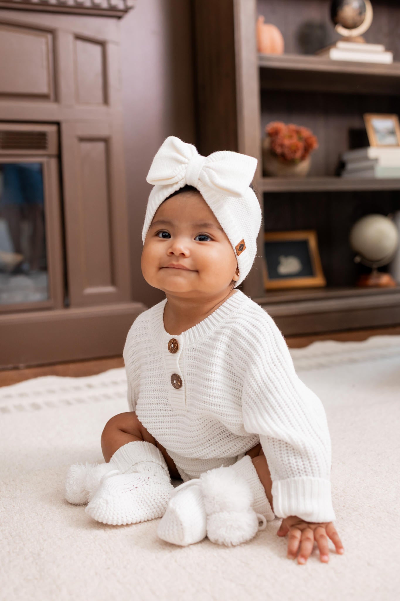 Baby sitting on a white carpet wearing Chunky Knit Romper, Chunky knit bow headband and knit booties in cloud