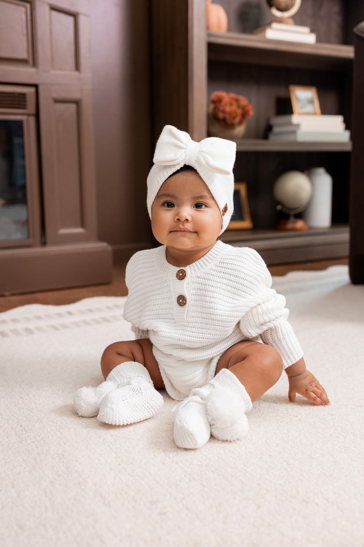 Baby sitting on a white carpet wearing Chunky Knit Romper, Chunky knit bow headband and knit booties in cloud