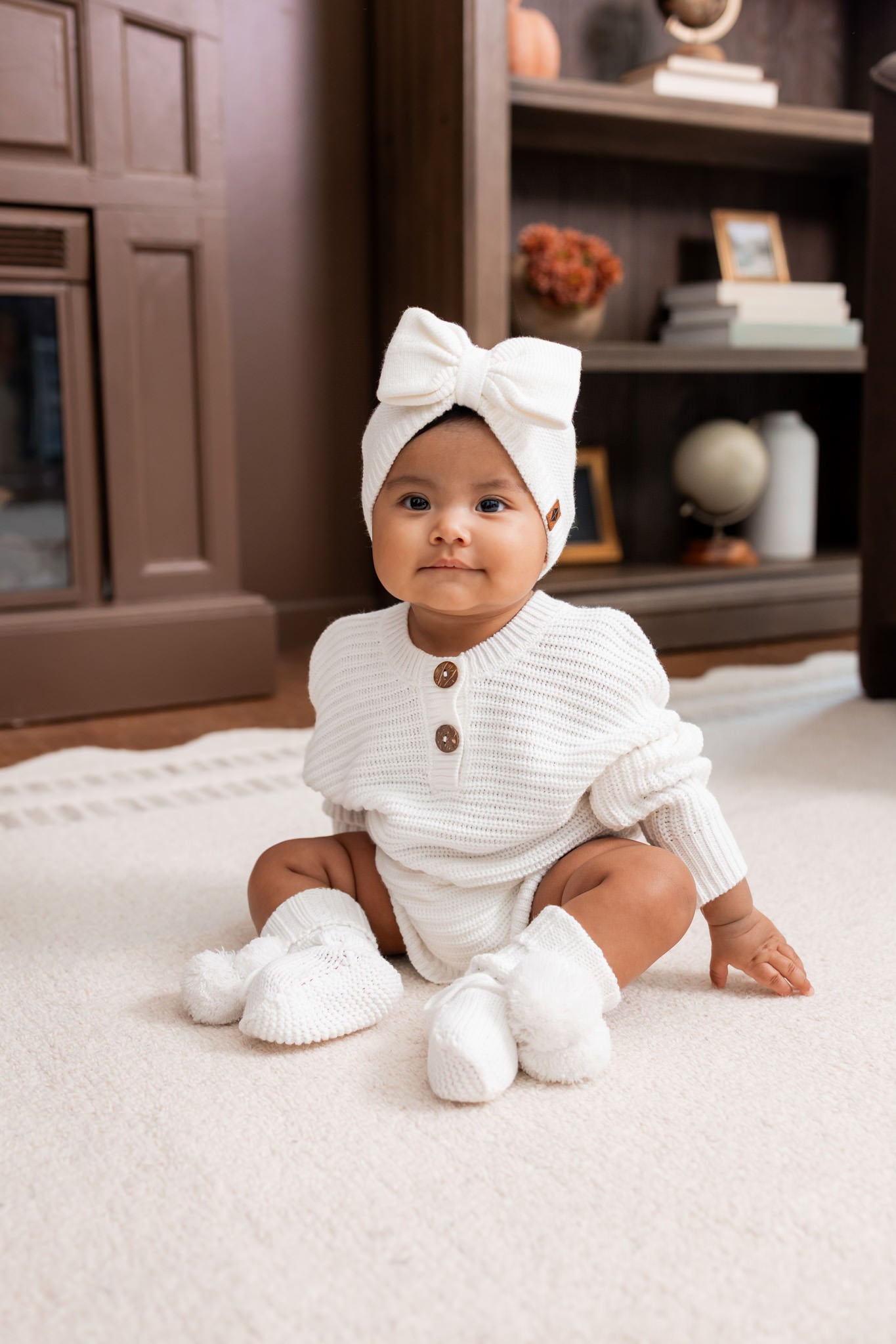Baby sitting on a white carpet wearing Chunky Knit Romper, Chunky knit bow headband and knit booties in cloud