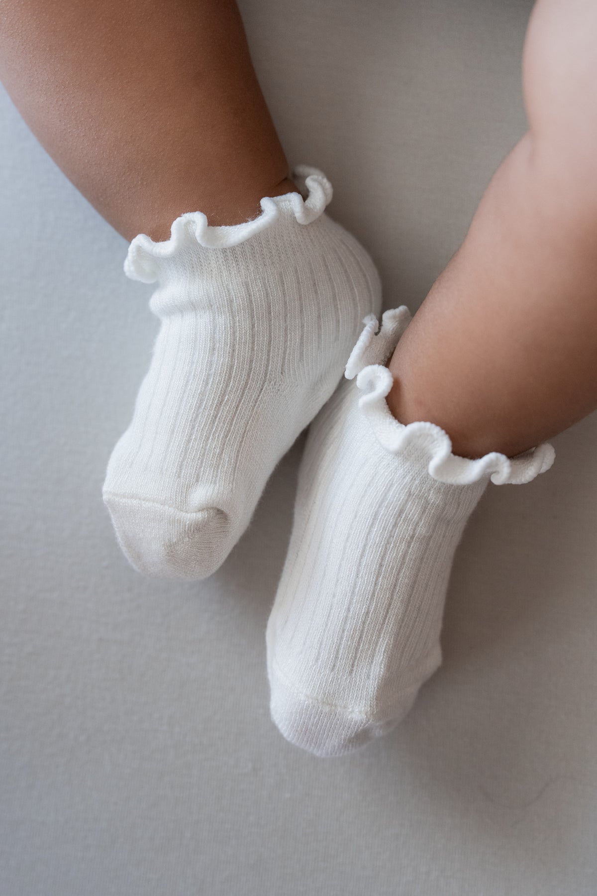 White ribbed ankle socks with ruffled trim worn on a baby's feet, photographed from above on a soft neutral surface.