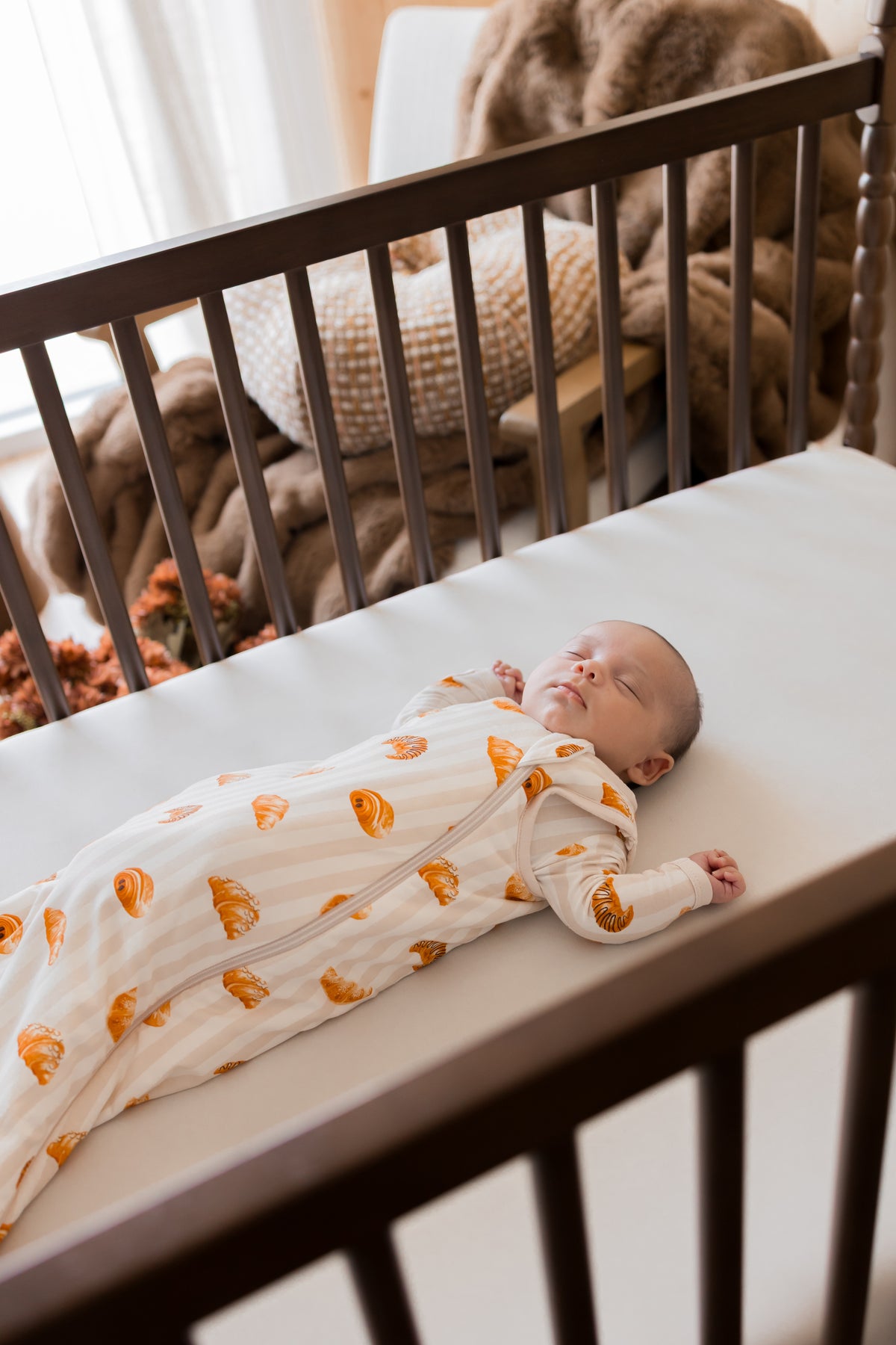 Baby sleeping in Sleep Bag with Croissant 1.0 print in a crib on a beige crib sheet.