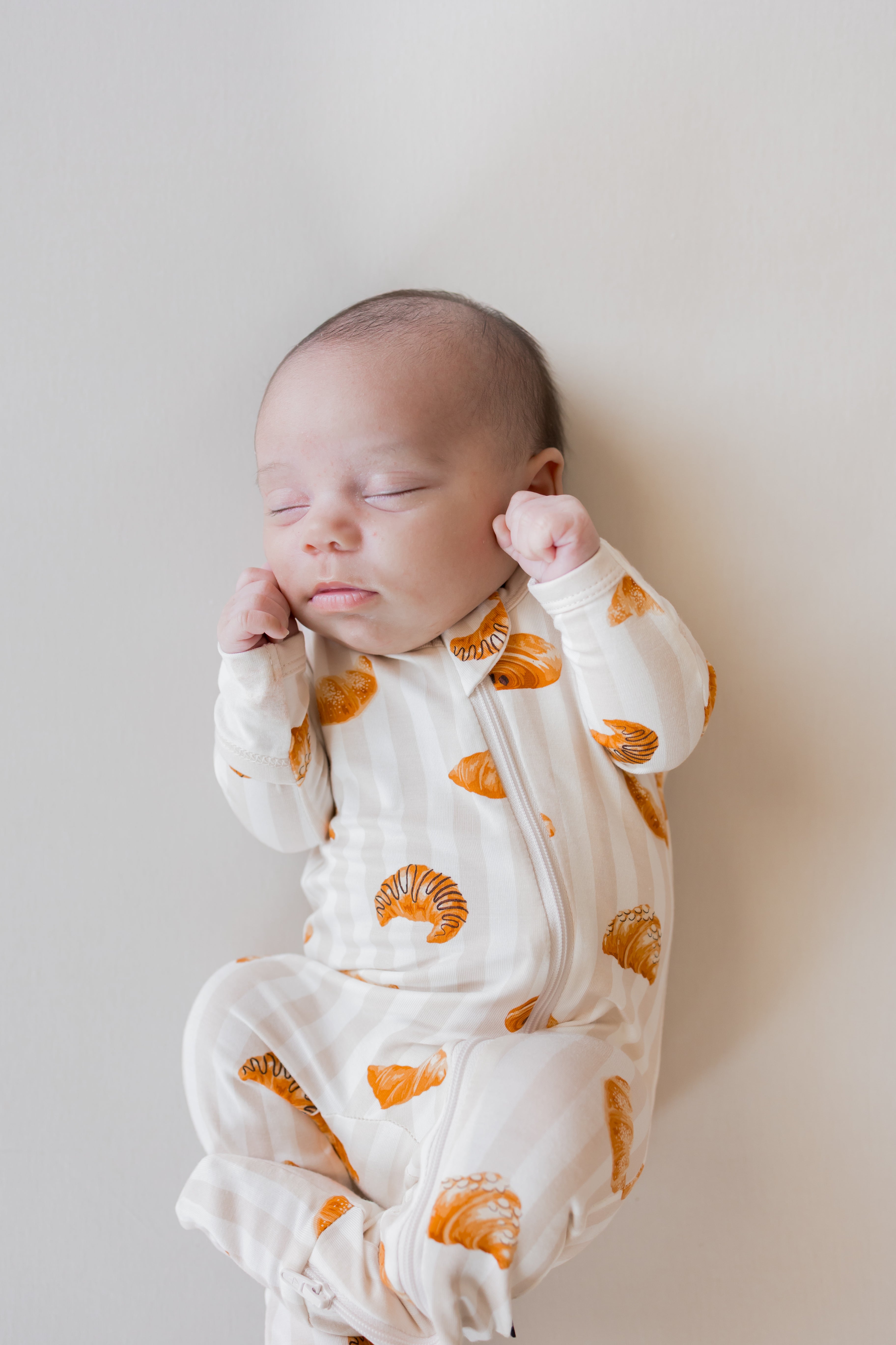 Baby sleeping in Zippered Footie with Croissant print on a beige background