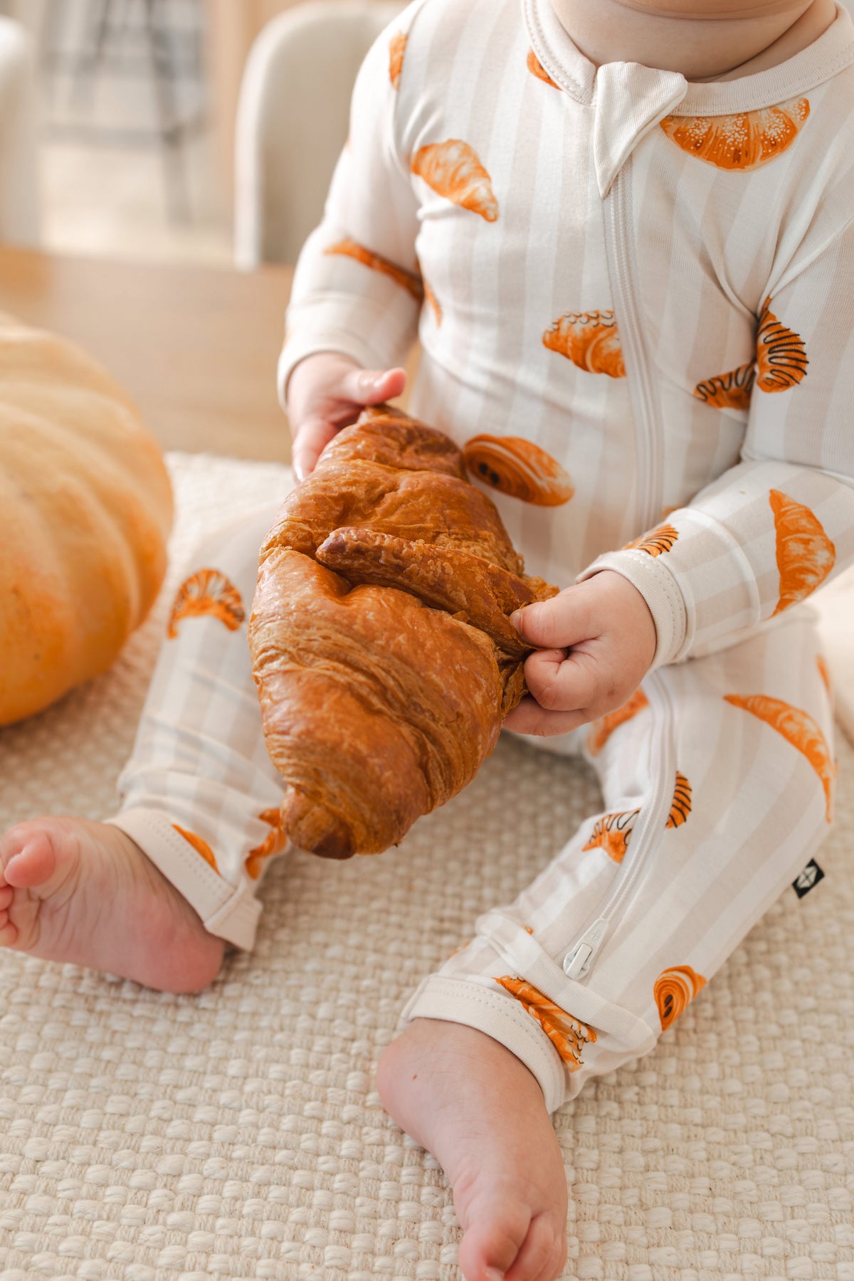 Close up of Baby holding a croissant while wearing Zippered Romper with Croissant print