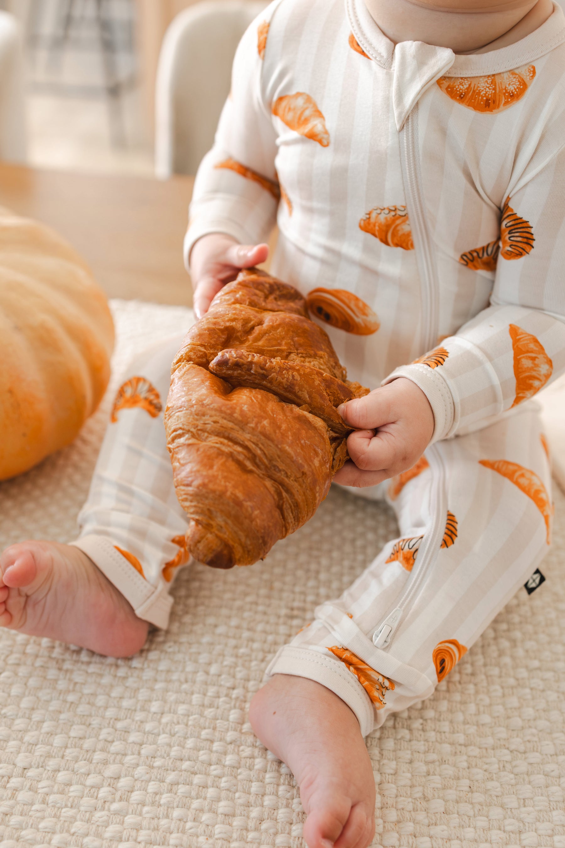 Close up of Baby holding a croissant while wearing Zippered Romper with Croissant print