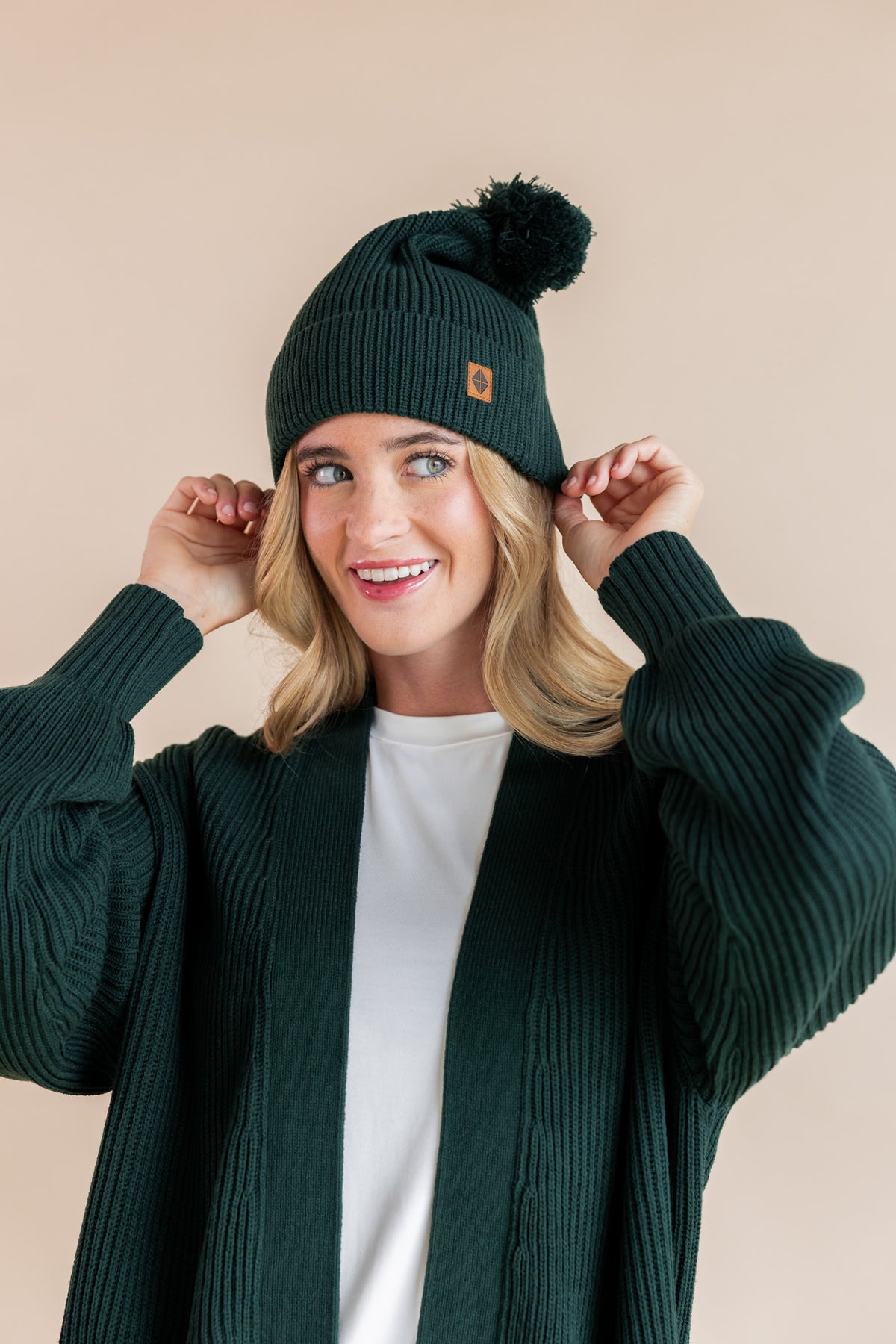 Modal smiling in chunky knit women's oversized cardigan and chunky knit adult ribbed beanie in cypress over white t-shirt on a beige background