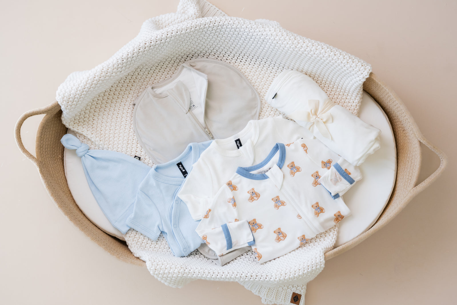 Baby clothes including take me home sets and footies in a basket on a beige background