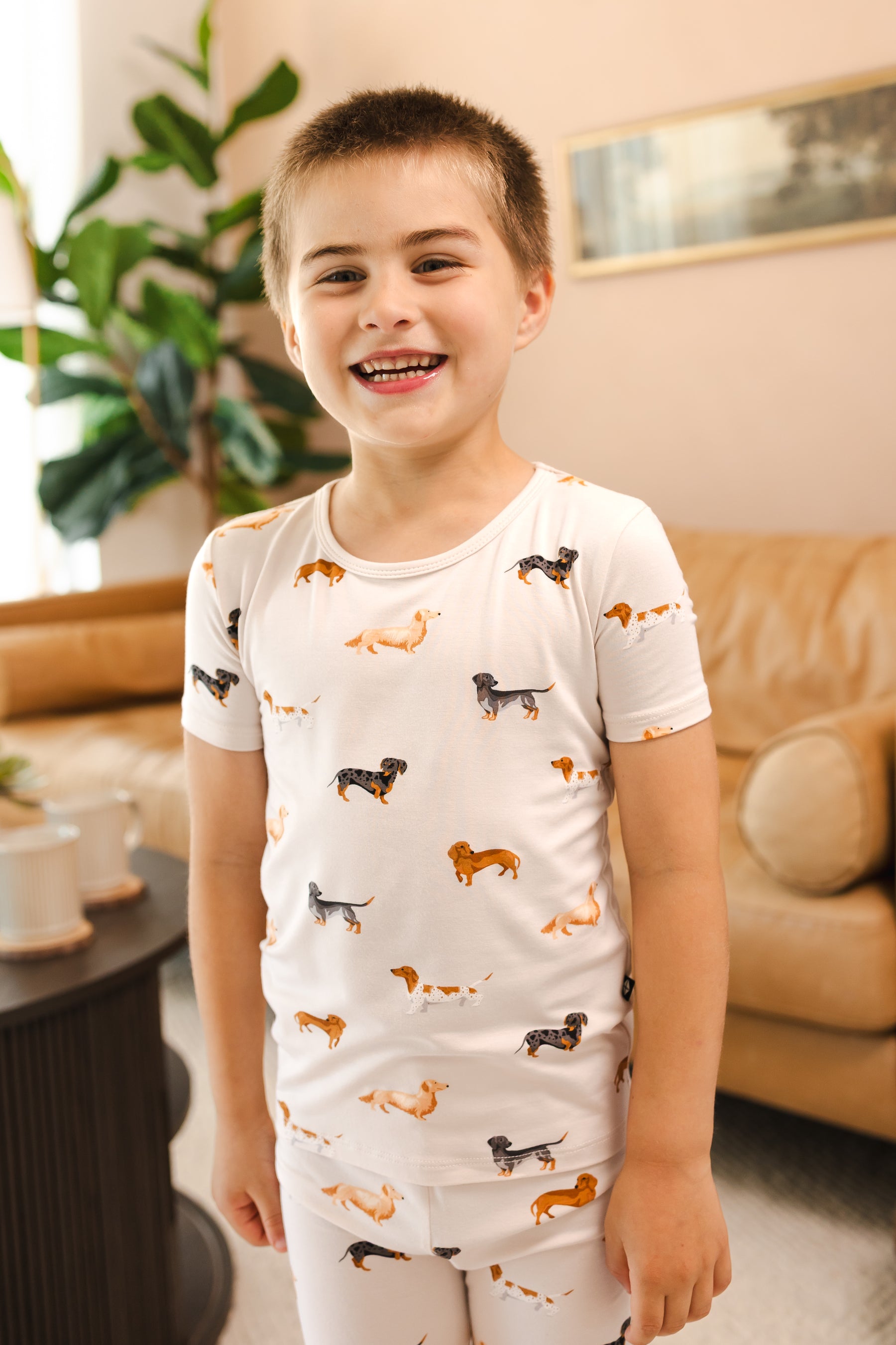 Boy smiling in beige Short Sleeve Pajamas in Dachshund print in a living room 