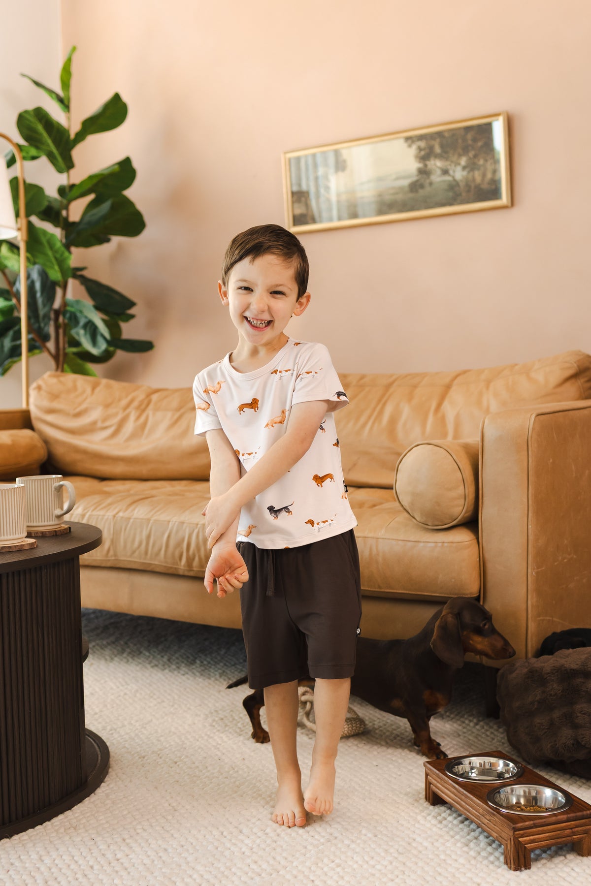 Boy standing in a living room smiling in a Beige Toddler Basic Tee in Dachshund print over brown Drawstring with a Dachshund dog.