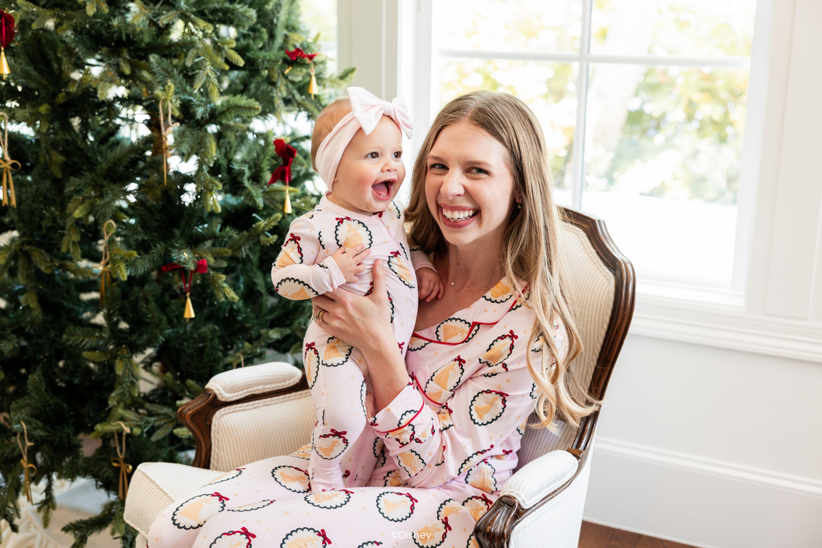 Woman and baby wearing matching pink pajamas with a disney princess pattern with a christmas tree in the background