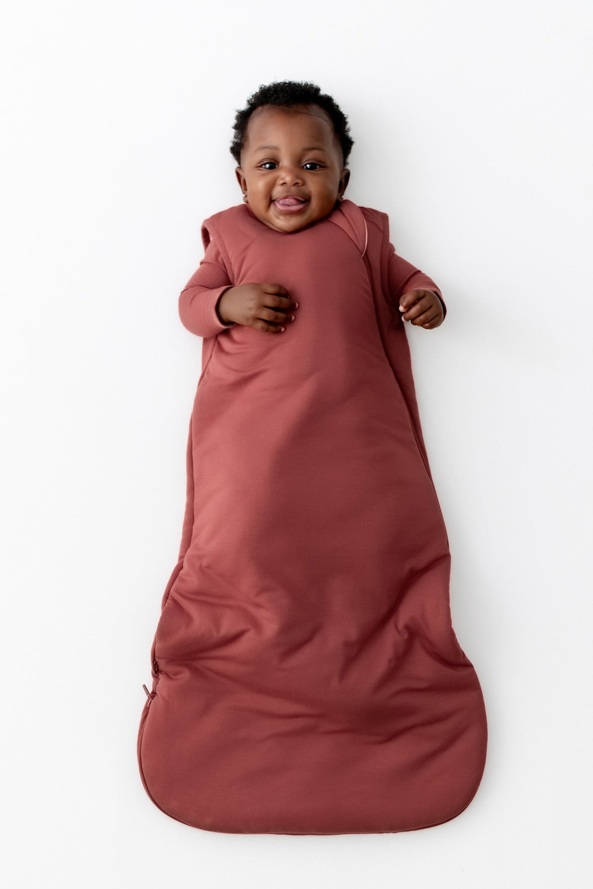 A baby lying on their back wrapped in a dusty terracotta sleeping bag, set against a plain white background.