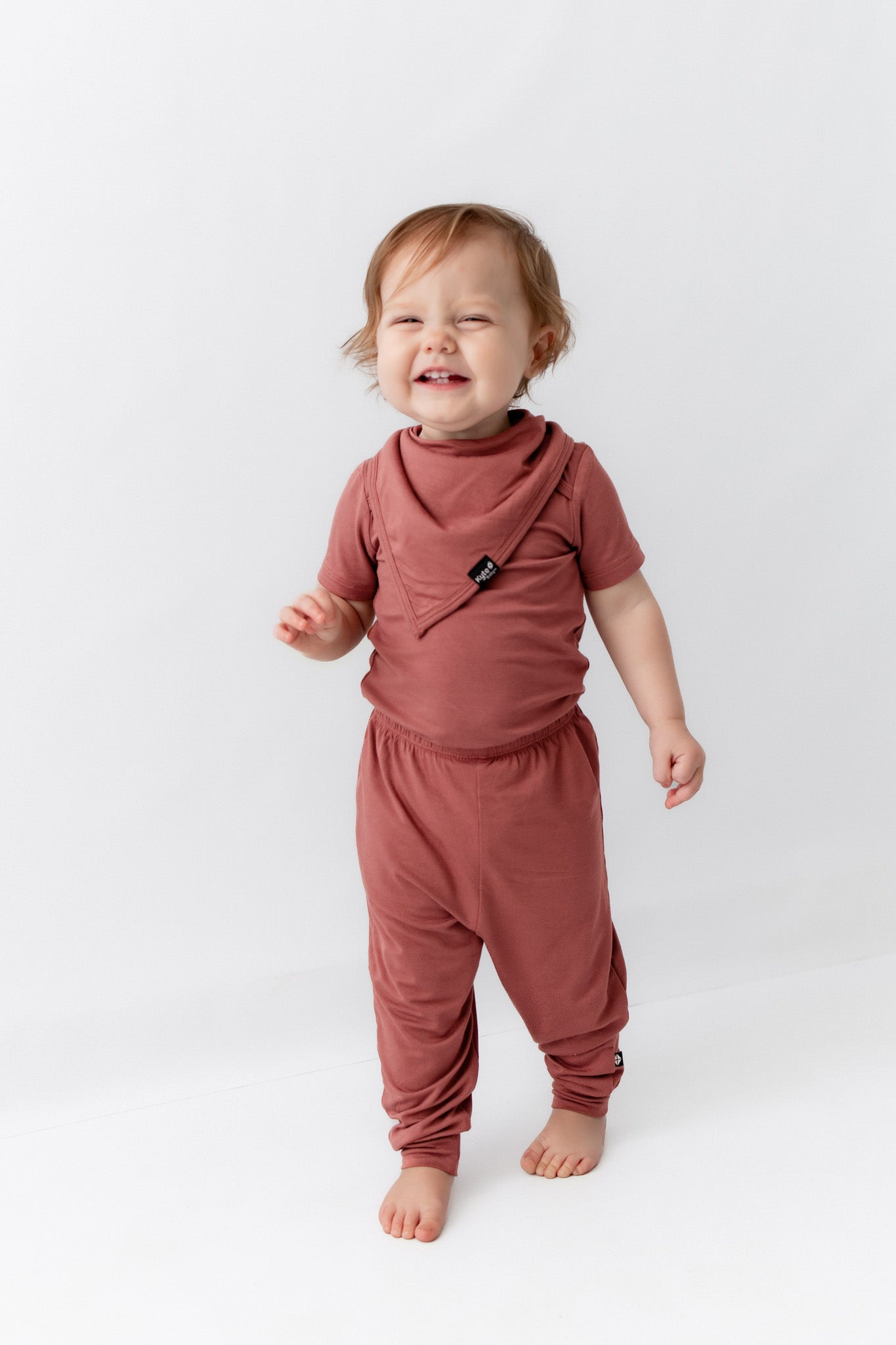 Child wearing Bib in Ember over similar color shirt and pant on a white background