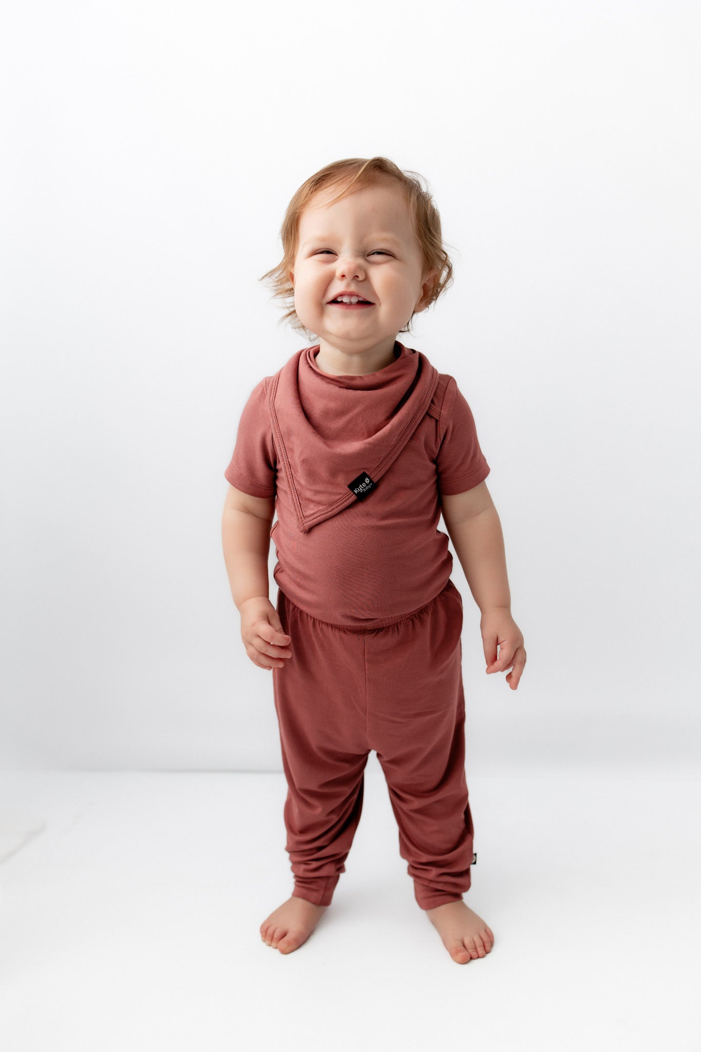 Child smiling while wearing Bib in Ember over similar color shirt and pant on a white background