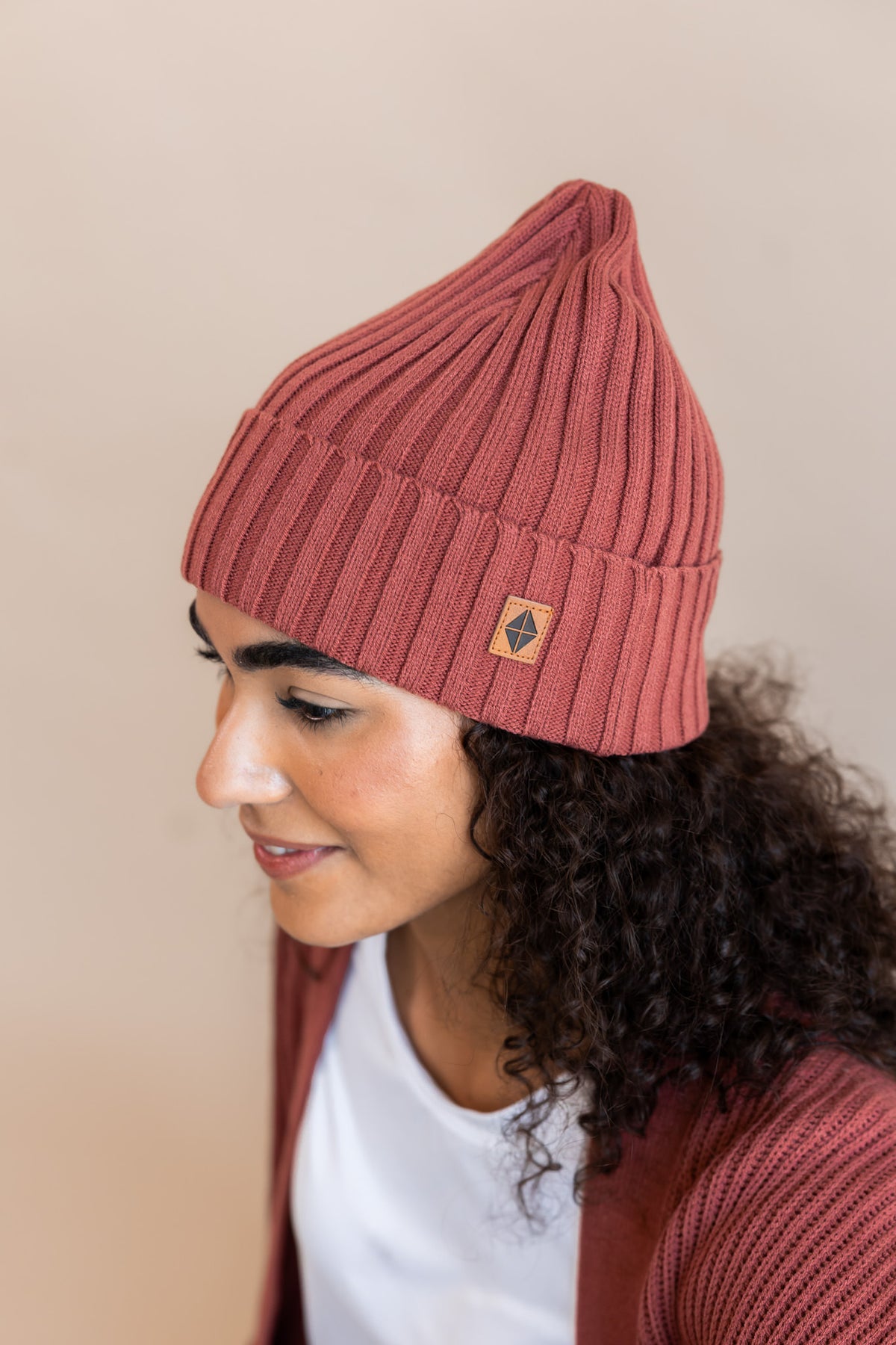 Side pose of Modal wearing Chunky Knit Adult Ribbed Beanie in Ember over white T-shirt and Chunky knit cardigan in ember on a beige background