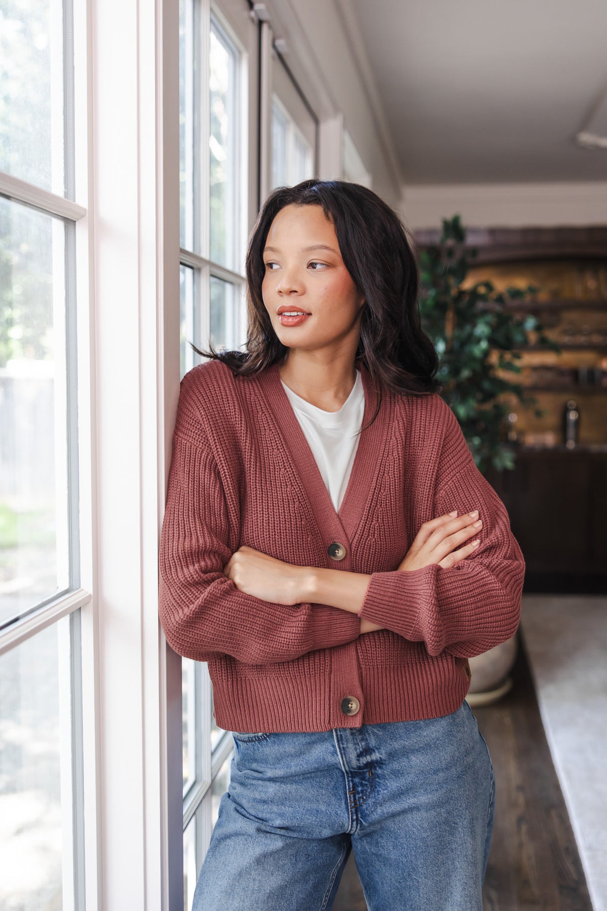 Modal standing against a window wearing Chunky Knit Women's Cropped Cardigan In Ember over white T-shirt and blue jeans