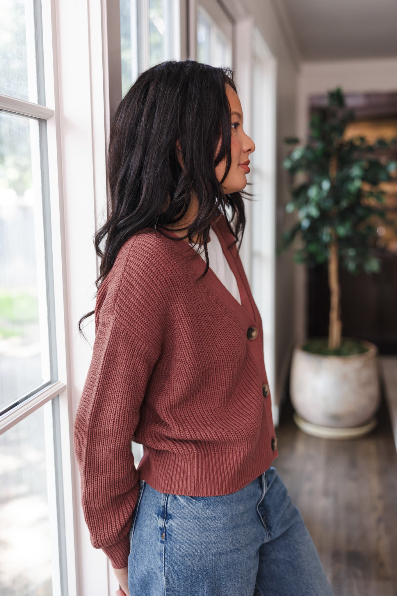 Modal standing against a window wearing Chunky Knit Women's Cropped Cardigan In Ember over white T-shirt and blue jeans