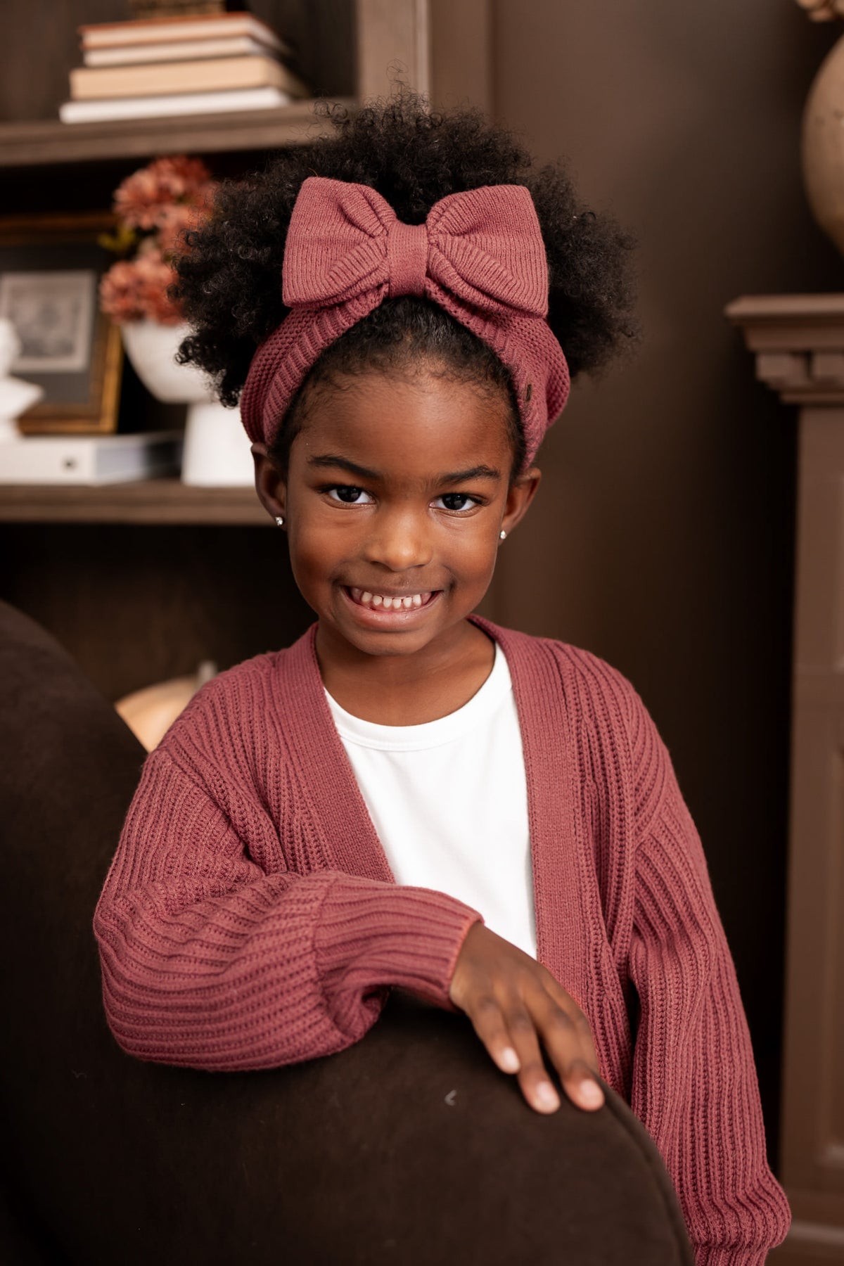 Child posing in living room wearing Chunky Knit Bow Headband in Ember over White T-shirt and chunky knit over sized cardigan in ember
