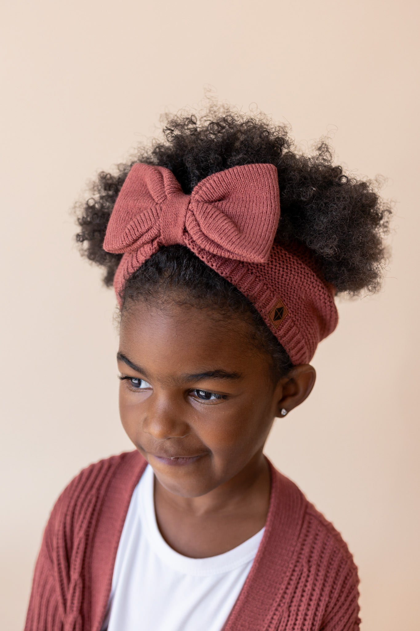 Child wearing Chunky Knit Bow Headband in Ember over White T-shirt and chunky knit oversized cardigan in ember on a beige background