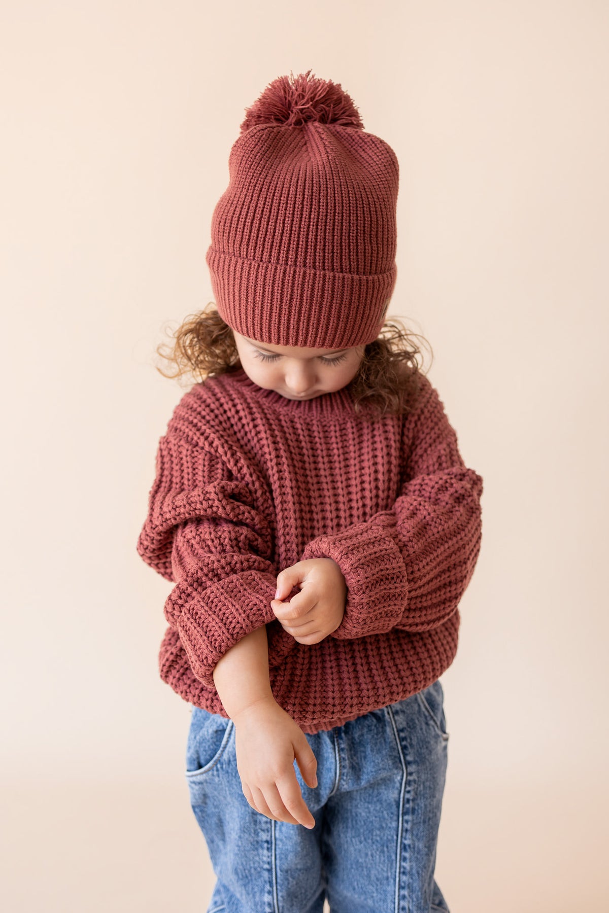 Girl adjusting the sleeves of Chunky Knit Pom Beanie in Ember over Chunky knit sweater in ember and blue jeans on a beige background