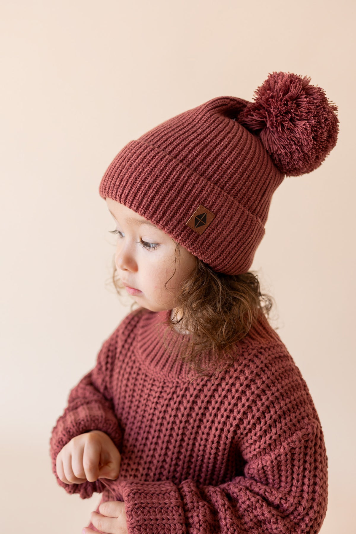 Girl wearing Chunky Knit Pom Beanie in Ember over Chunky knit sweater in ember on a beige background