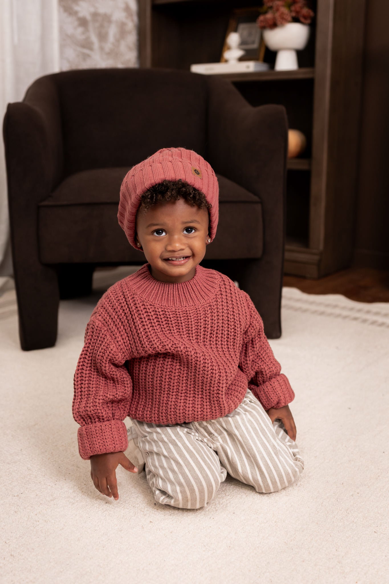 Boy wearing Chunky Knit Ribbed Beanie and chunky knit sweater in Ember over beige pants sitting on white carpet