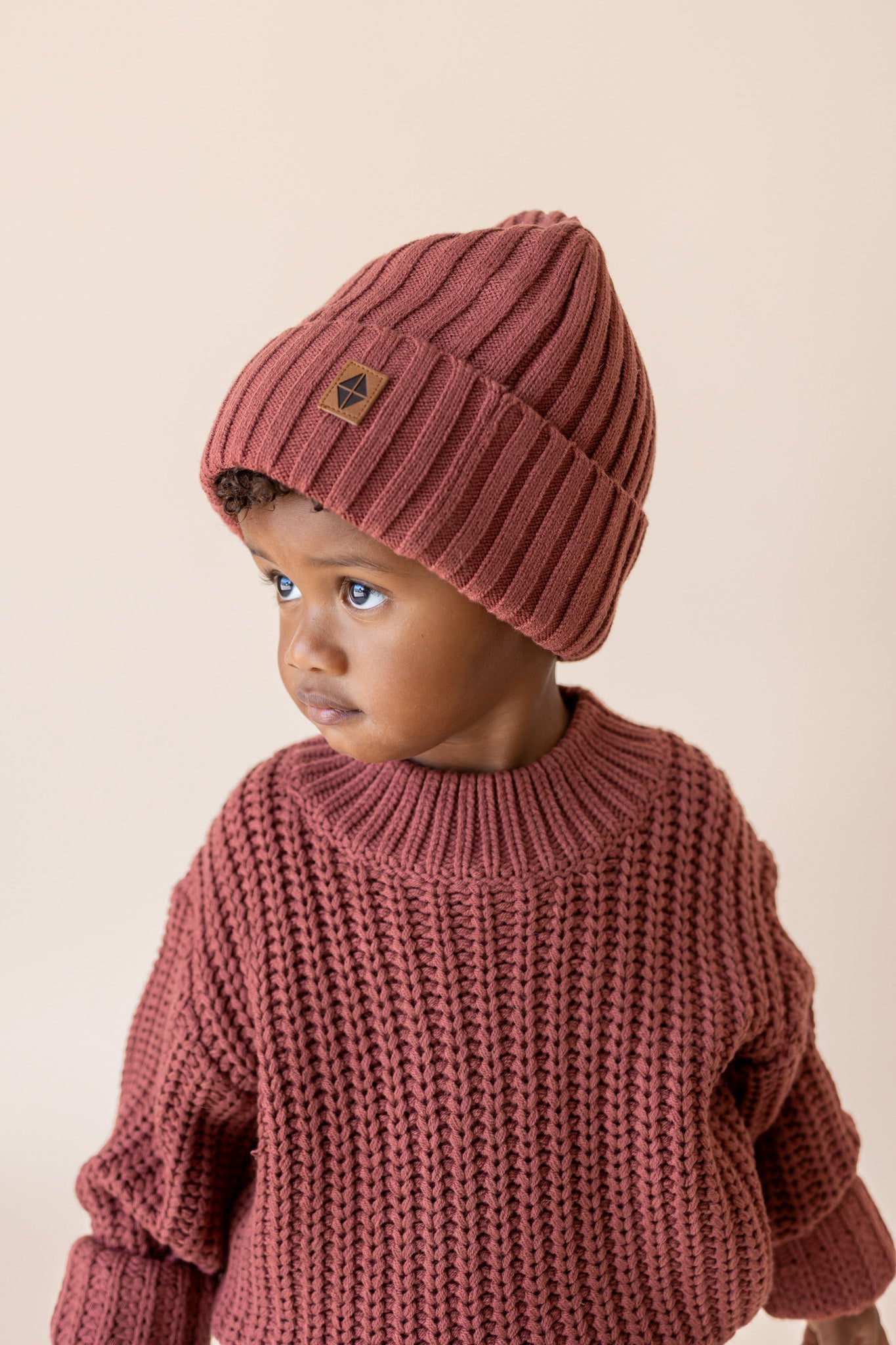 Boy wearing Chunky Knit Ribbed Beanie and chunky knit sweater in Ember on a beige background