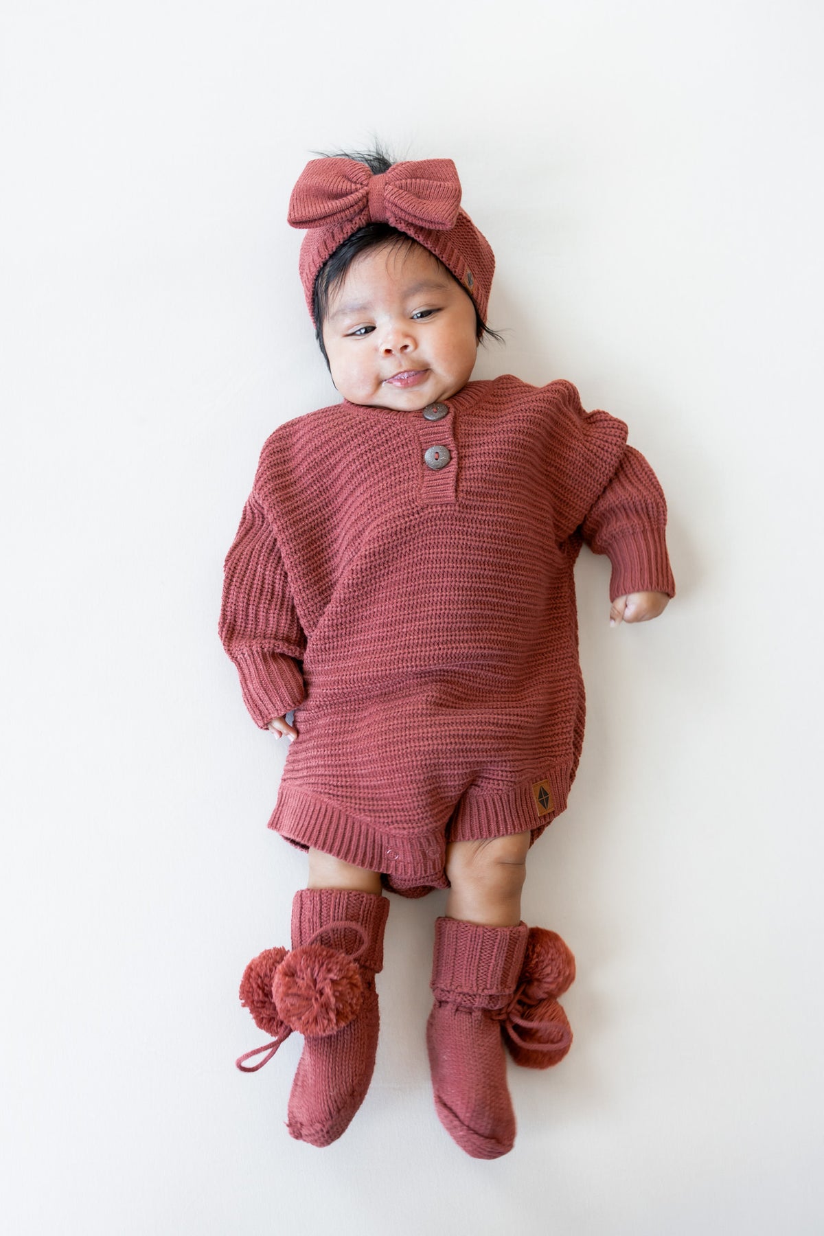 Baby wearing Chunky Knit Romper, chunky knit bow headband and knit booties in Ember lying on a white background