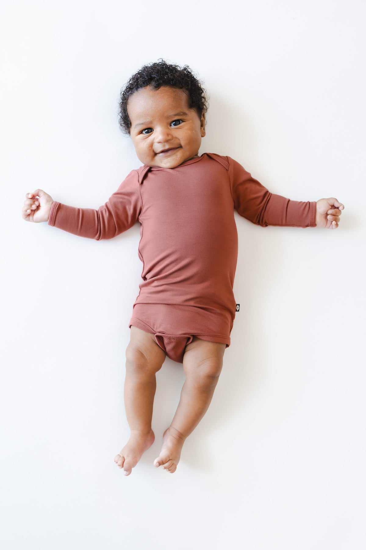 A smiling baby lying on their back wearing a terracotta-colored long-sleeve bodysuit against a clean white background.