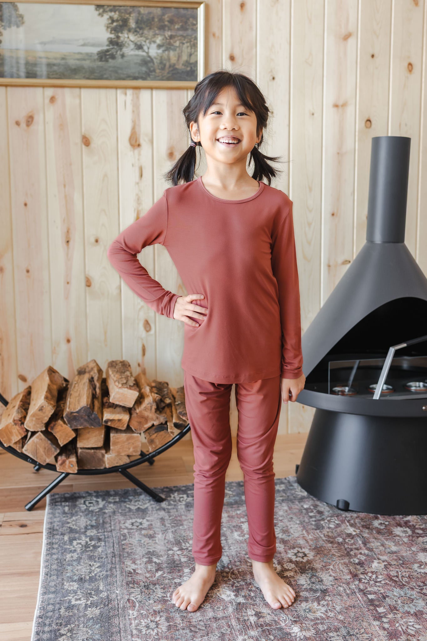 A young girl stands barefoot in a cozy room with wood-paneled walls, wearing a matching rust-colored long-sleeve top and pants pajama set. A black fireplace is to her right, and a stack of firewood sits on a black rack to her left.