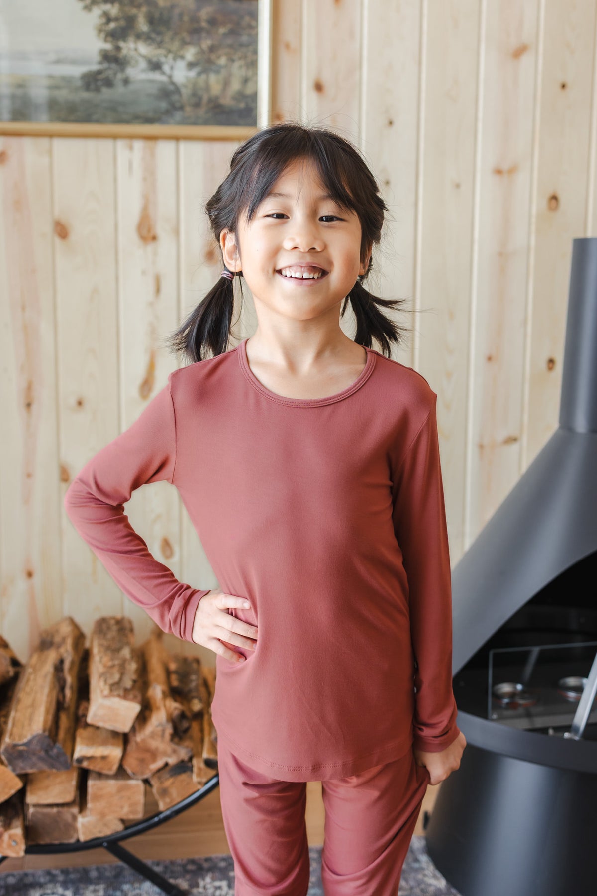 A young girl with dark hair in pigtails stands smiling in a cozy room with light wood-paneled walls, wearing a matching dusty terracotta long-sleeve top and pants pajama set.