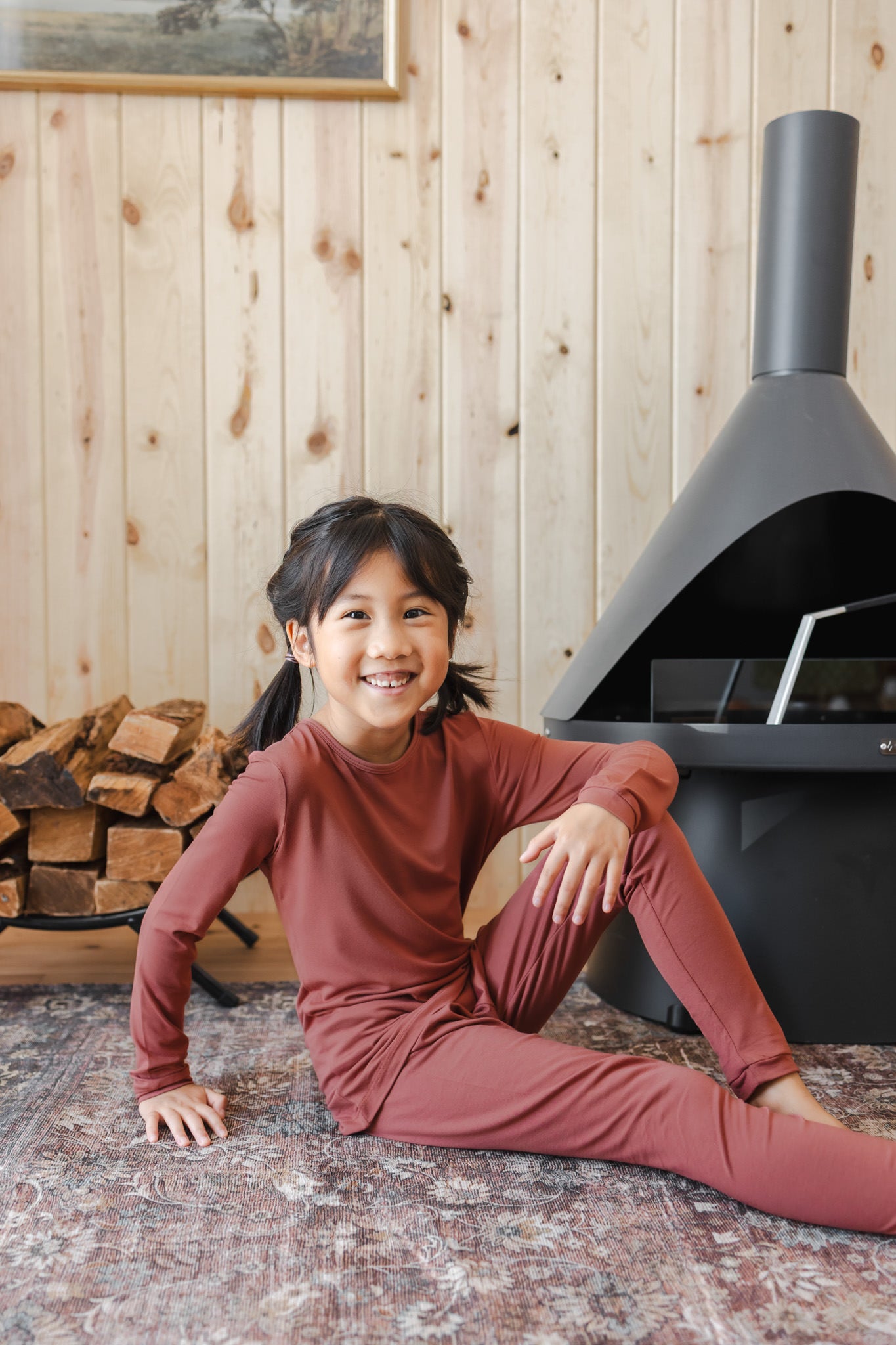 A smiling young girl sits on a patterned rug in front of a wood-burning stove, wearing a matching rust-brown long-sleeve top and pants pajama set.