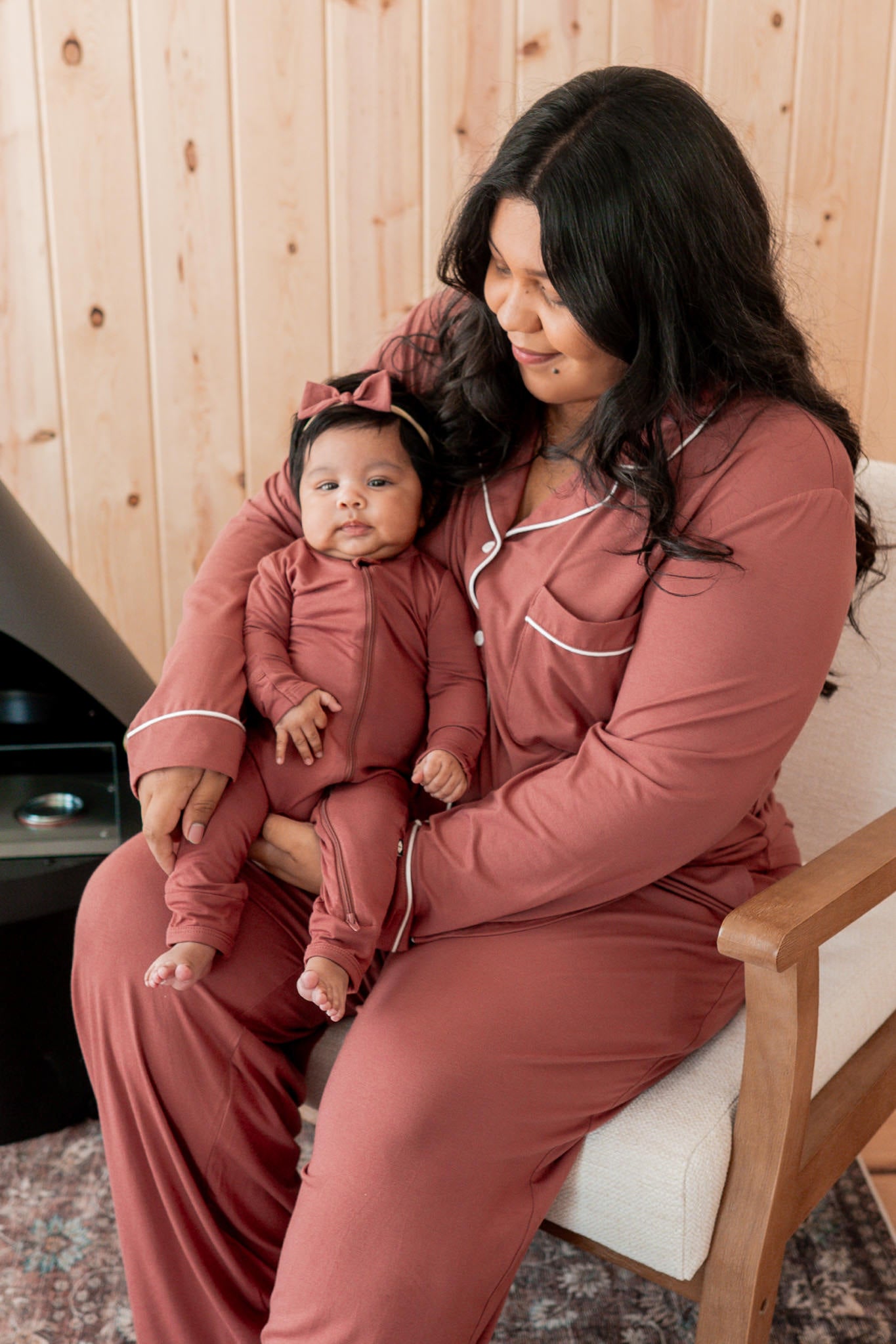 A woman and a baby wearing matching terracotta pajama sets sit together on a light-colored armchair in front of a wood-paneled wall.