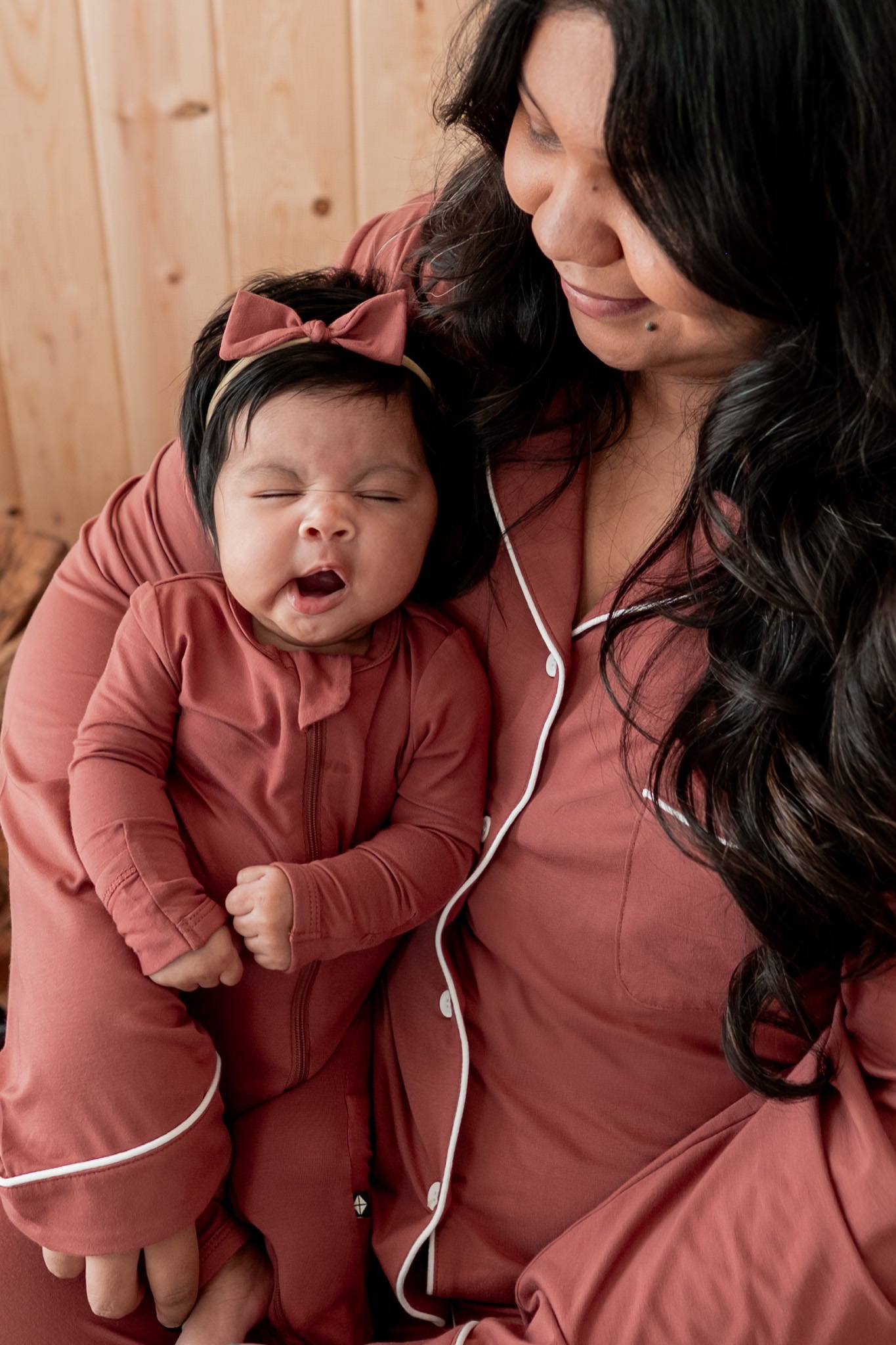 A woman and baby wearing matching terracotta pajamas, with the woman holding the baby who is wearing a matching long-sleeve romper and bow headband.