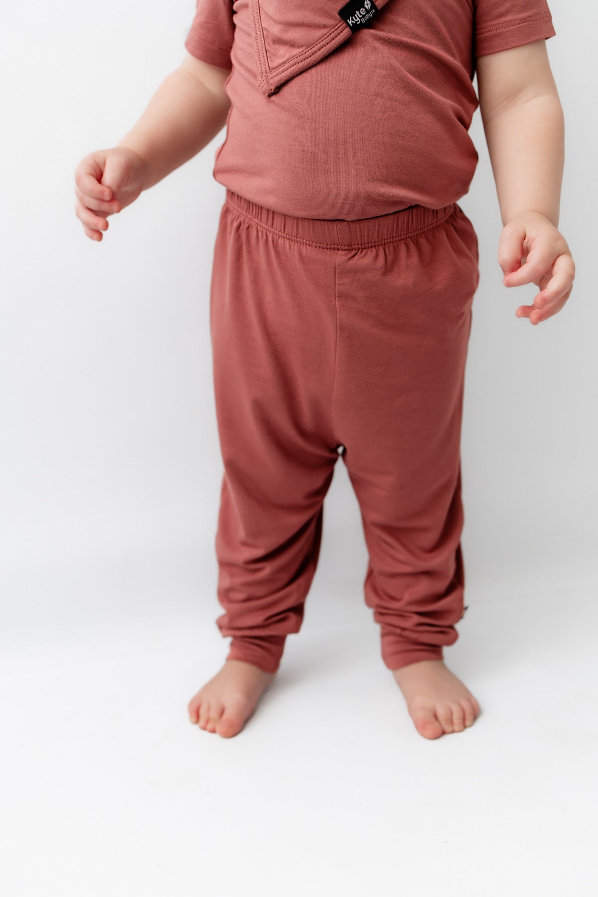 Child wearing Pant in Ember under a similar color shirt and bib on a white background